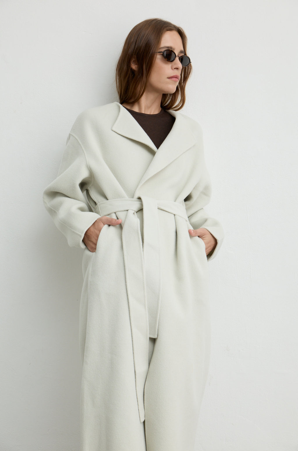 Woman wearing a light grey long coat with a tie belt, black sunglasses, and a brown t shirt against a light grey wall