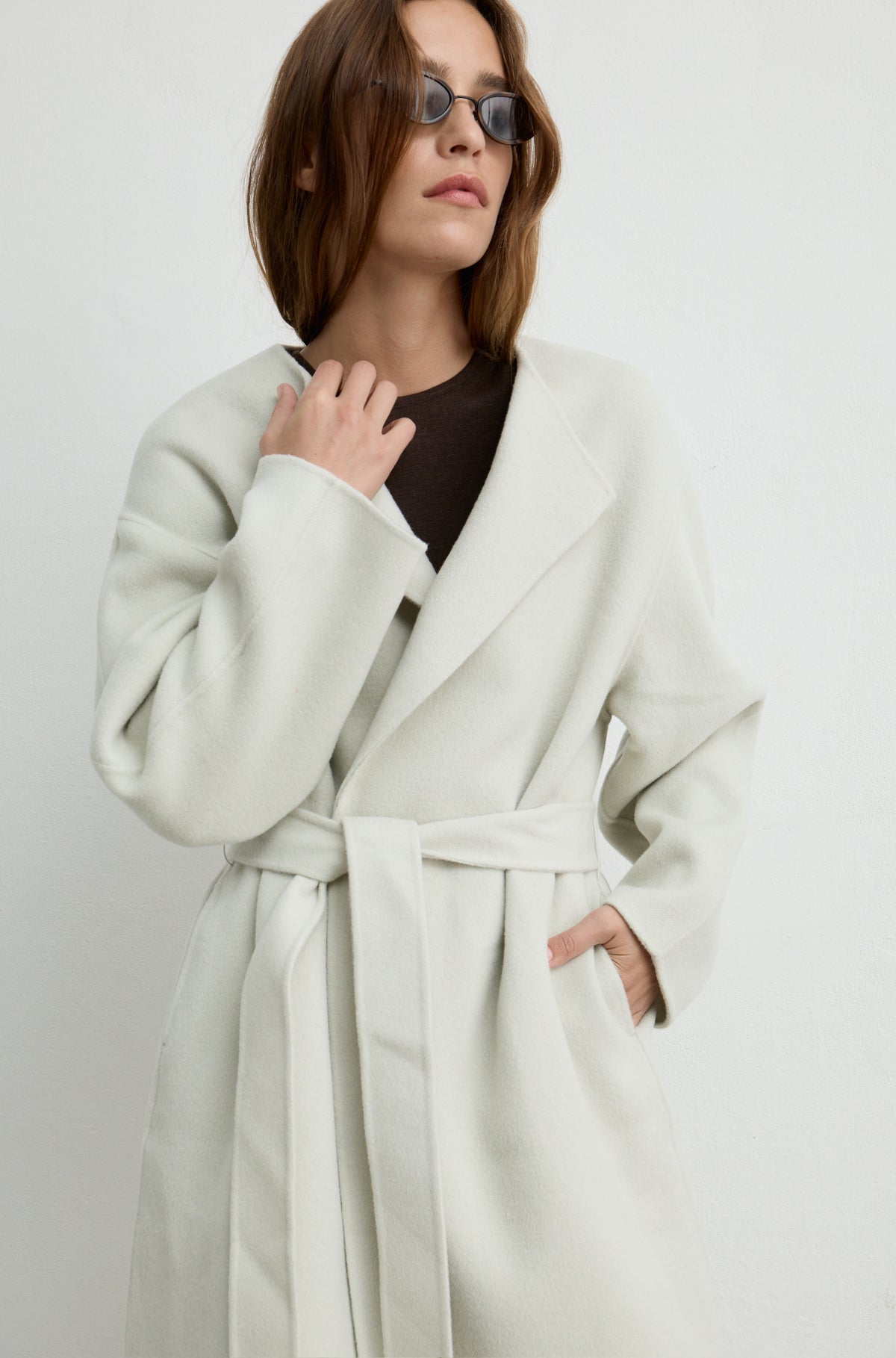 Woman wearing a light grey long coat with a tie belt, black sunglasses, and a brown t shirt against a light grey wall