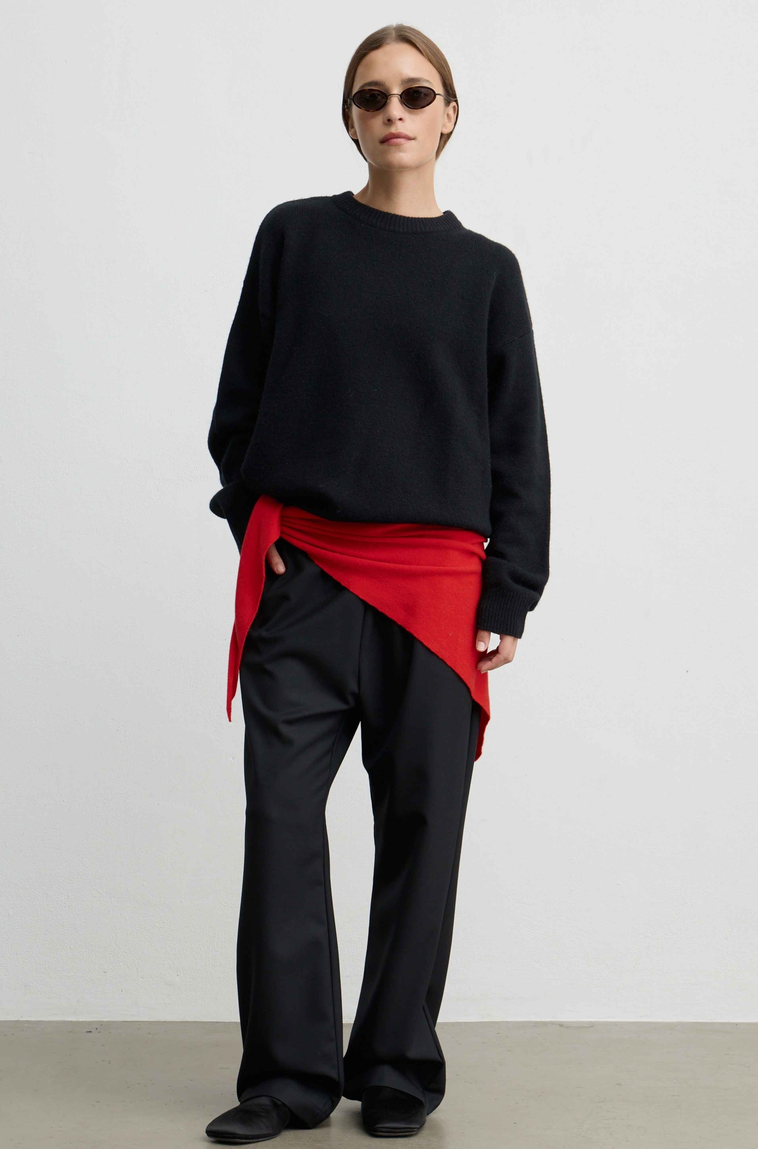 A model stands against a plain light gray studio wall wearing a black crewneck sweater, black wide-leg trousers, and a bright red asymmetrical wrap tied around the waist, along with dark sunglasses.