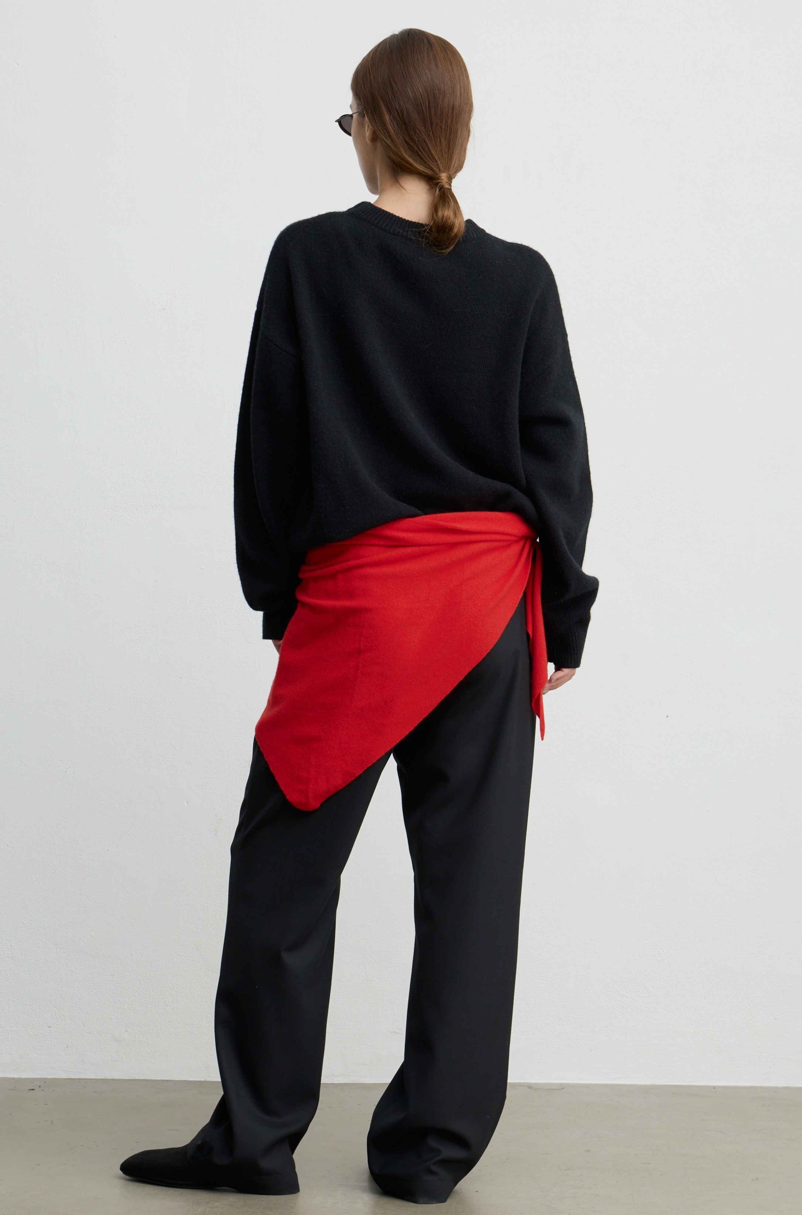 A model shown from the back wearing a black crewneck sweatshirt, black wide-leg trousers, and a bright red wrap-style shirt tied around the waist, set against a plain light gray studio background.