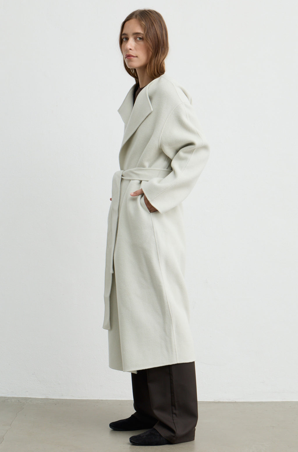 Woman wearing a light grey long coat with a tie belt, brown pants, and a brown t shirt against a light grey wall