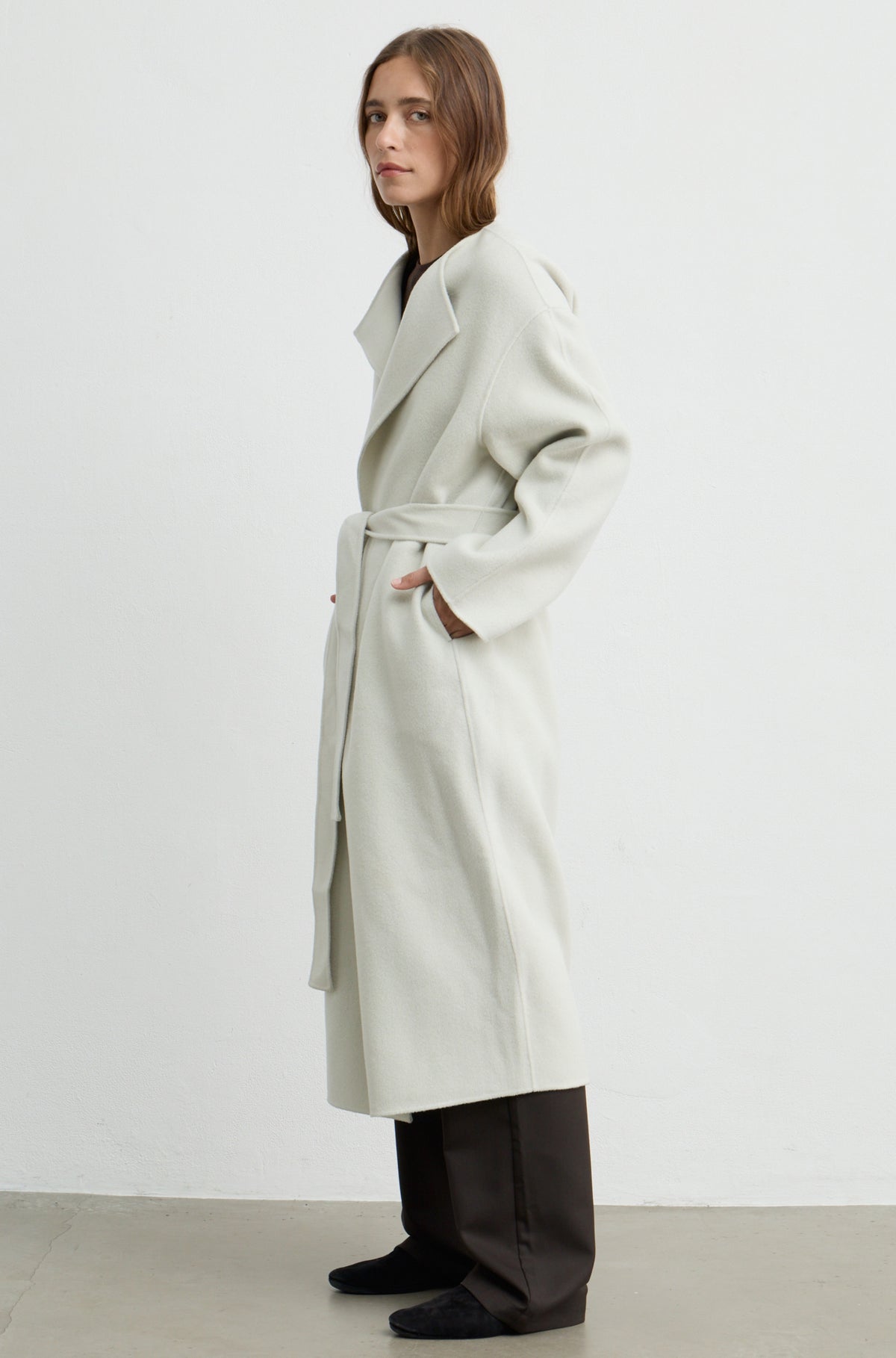 Woman wearing a light grey long coat with a tie belt, brown pants, and a brown t shirt against a light grey wall