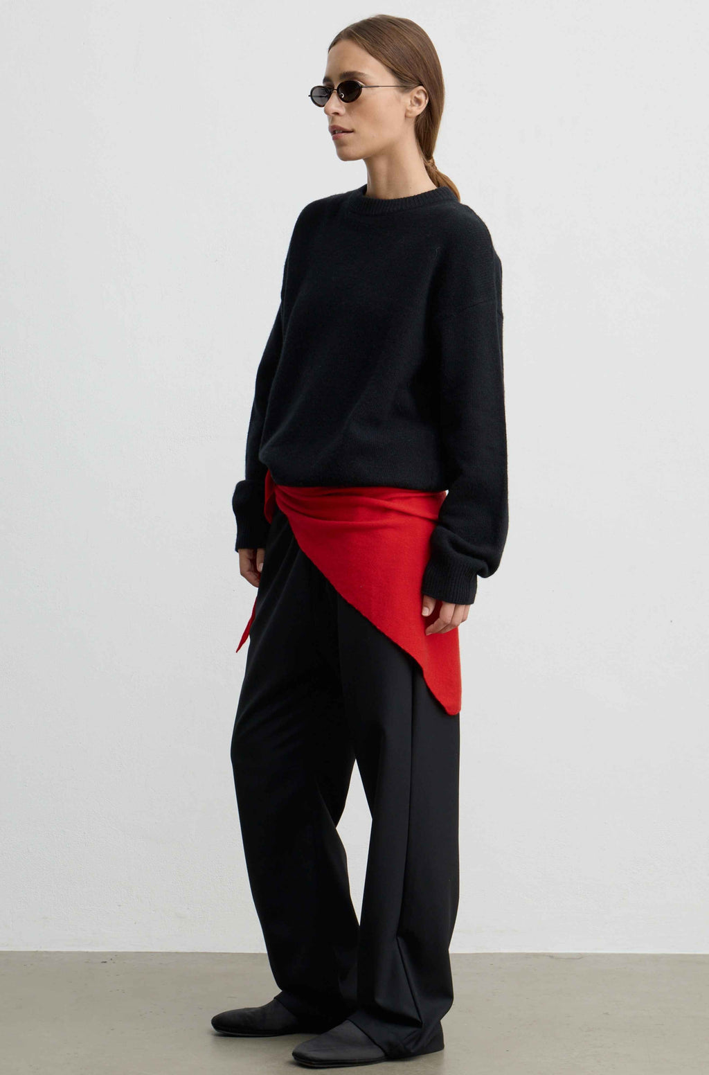 A model wearing a black crewneck sweater, black wide-leg trousers, and a bright red asymmetrical wrap tied at the waist, standing against a plain light gray studio background.