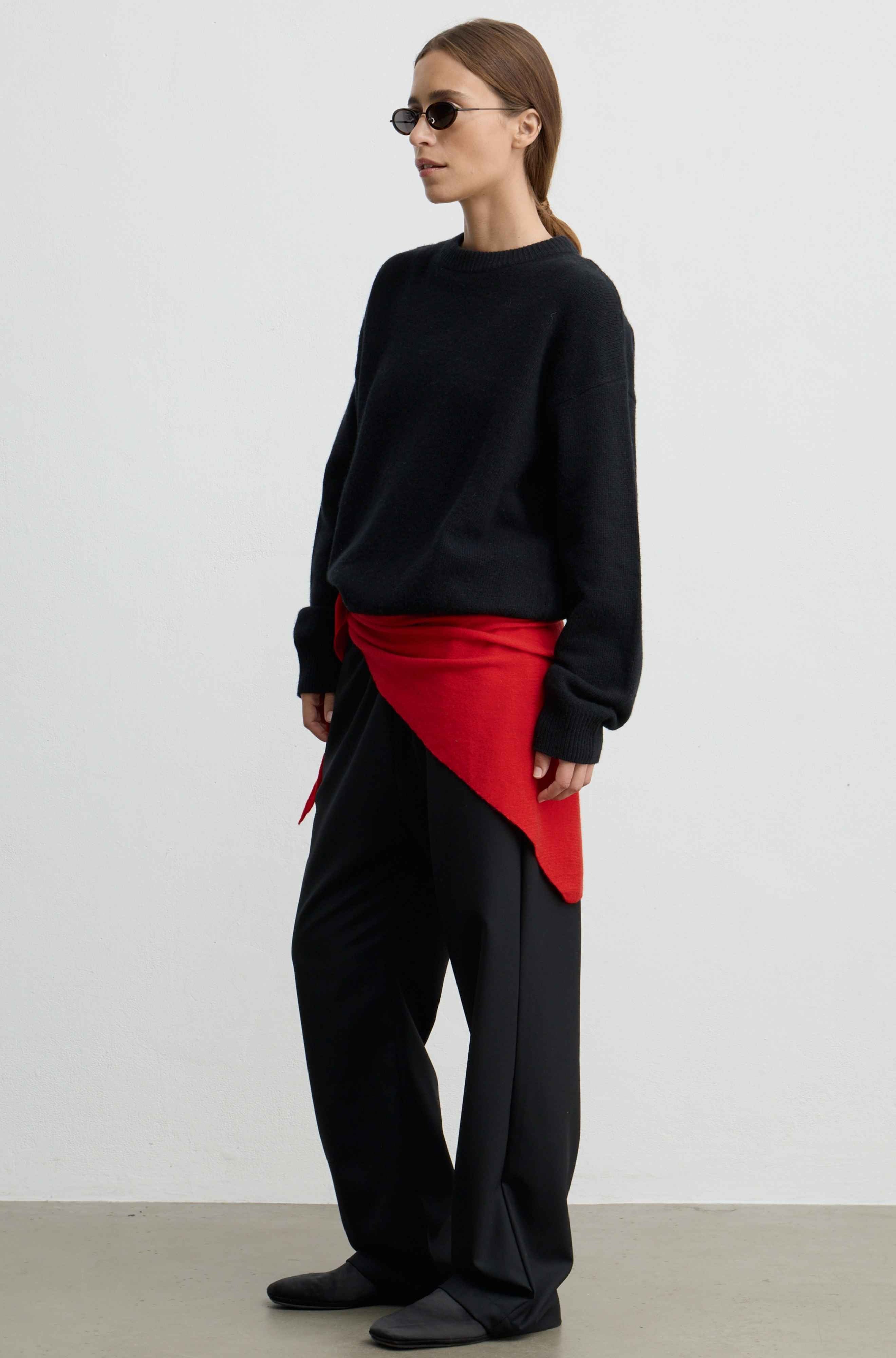 A model wearing a black crewneck sweater, black wide-leg trousers, and a bright red asymmetrical wrap tied at the waist, standing against a plain light gray studio background.