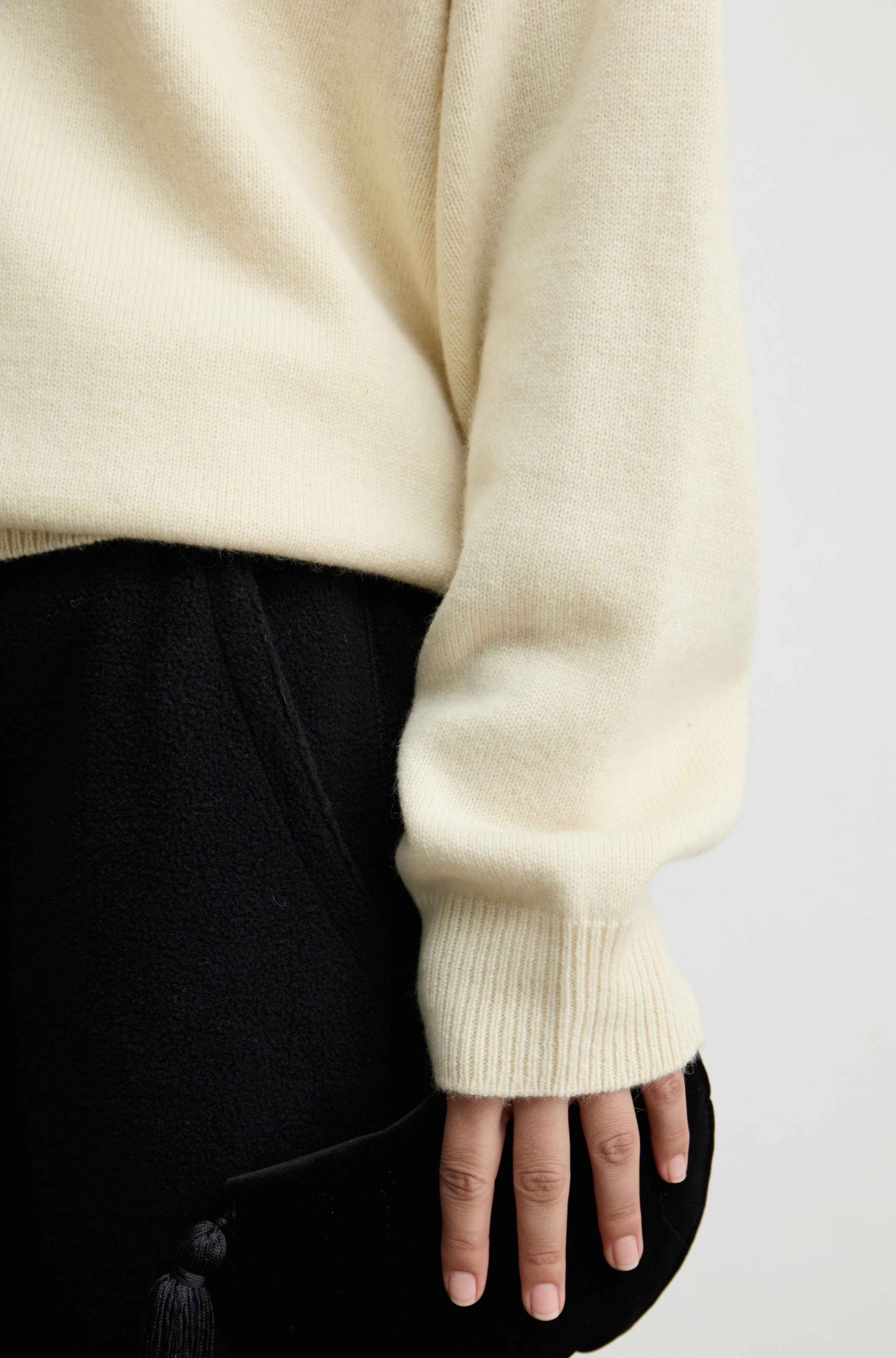 Close-up of a person wearing a cream-colored knit sweater with a ribbed cuff, paired with black bottoms, against a plain light background.