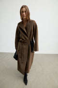 Woman wearing a dark brown wool wrap coat with a tie belt, black heels, and holding a black bag against a white wall