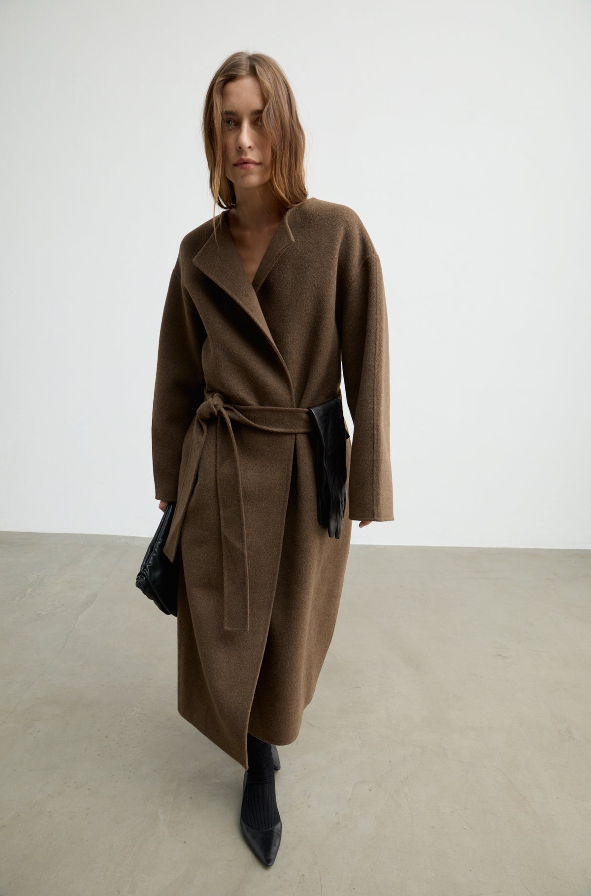 Woman wearing a dark brown wool wrap coat with a tie belt, black heels, and holding a black bag against a white wall