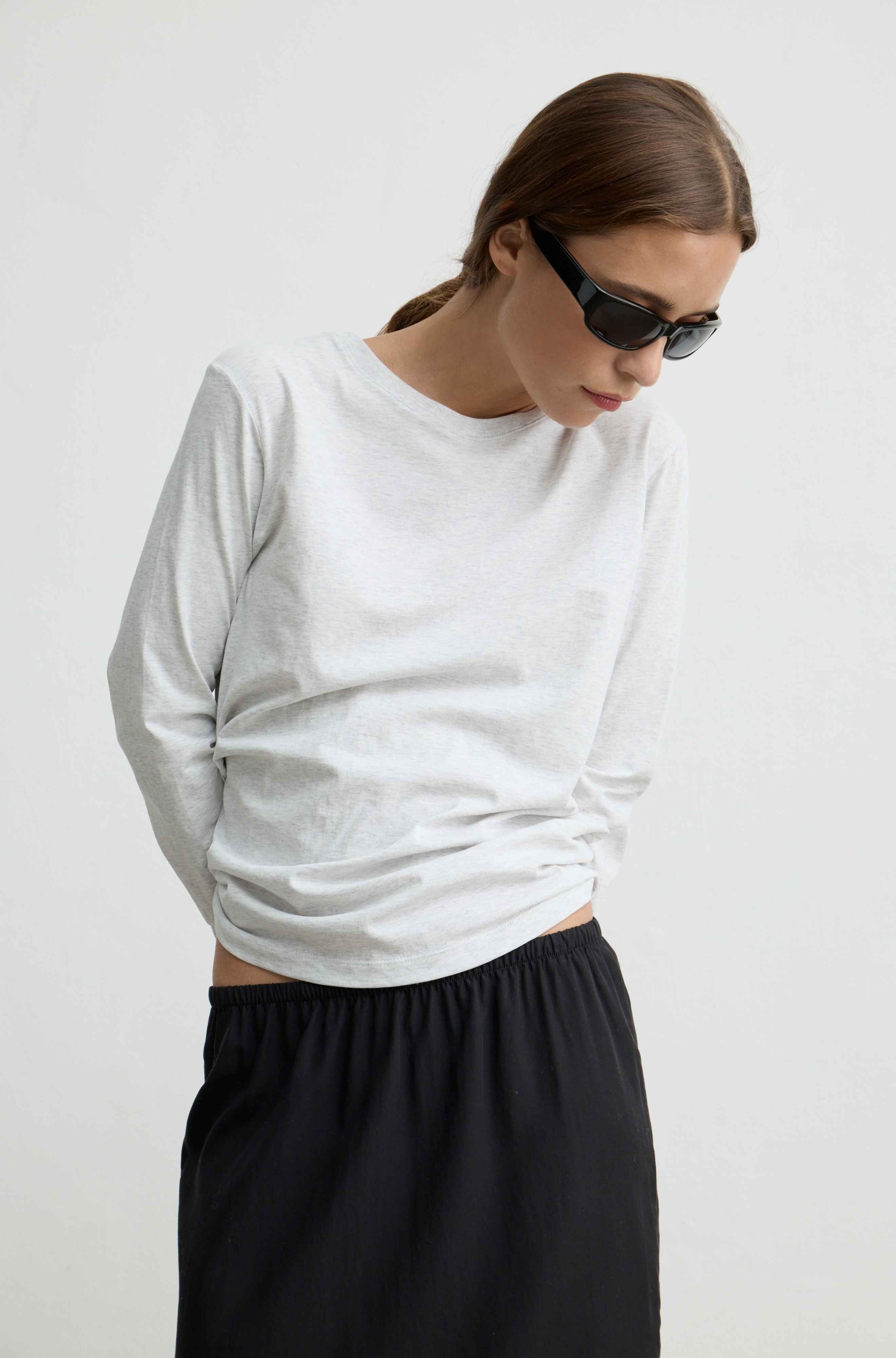 A woman wearing a light gray long-sleeve crewneck t-shirt and black pants, photographed against a plain light gray background.