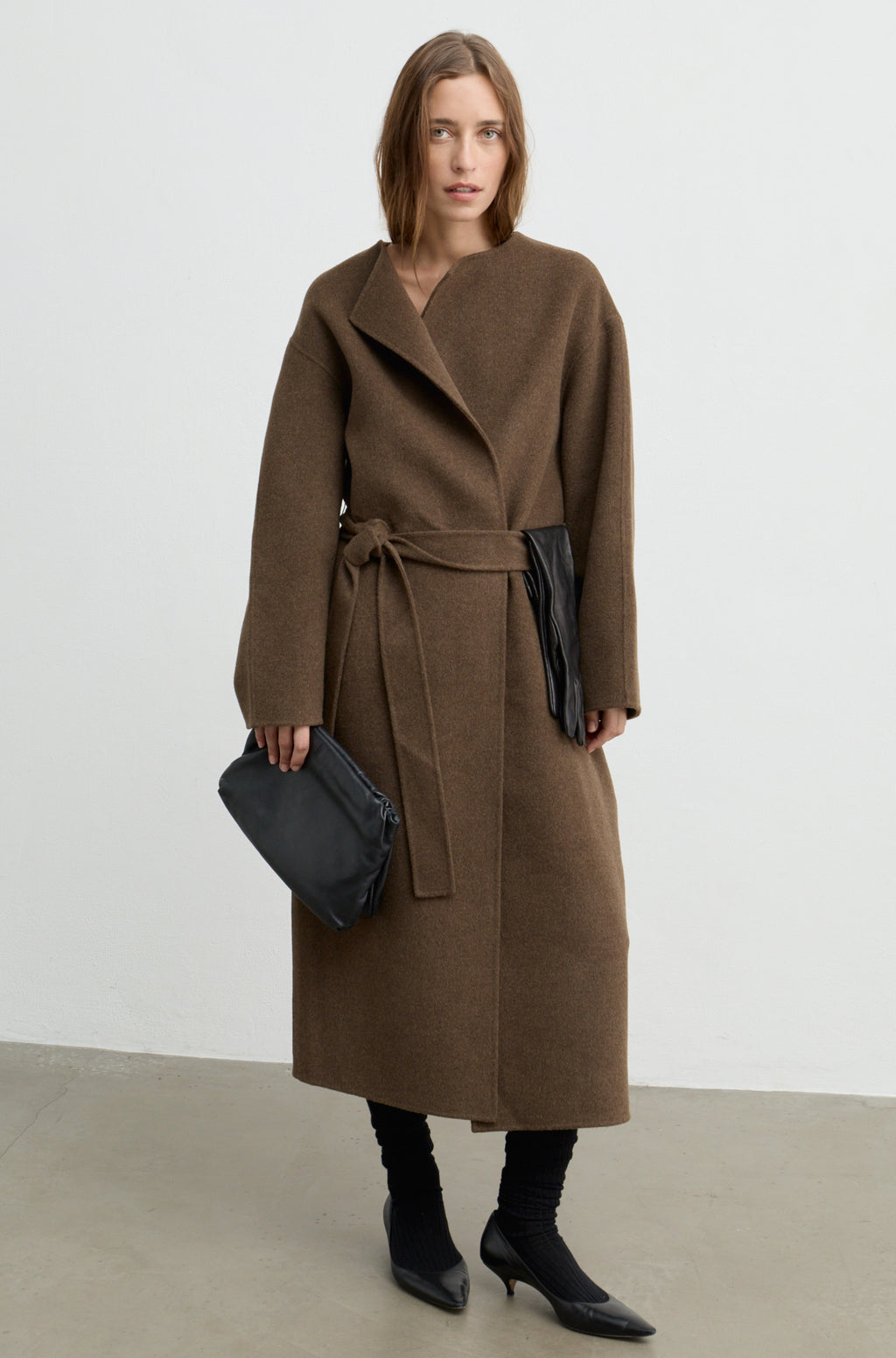 Woman wearing a dark brown wool wrap coat with a tie belt, black heels, and holding a black bag against a white wall