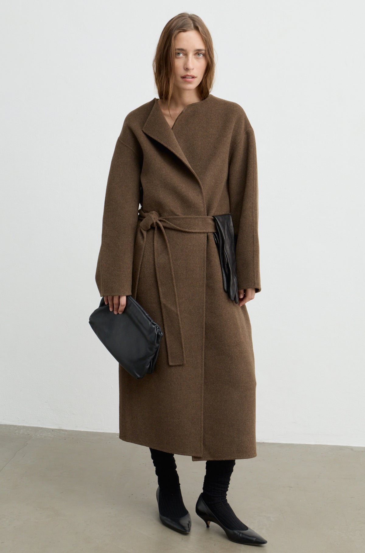 Woman wearing a dark brown wool wrap coat with a tie belt, black heels, and holding a black bag against a white wall
