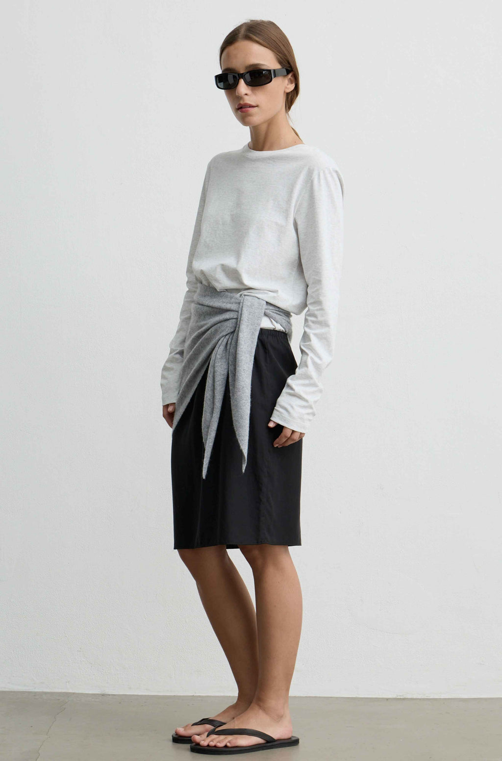 A model wearing a light gray long-sleeve top with a gray knit tie-waist sash, a black midi skirt, and black flip-flops, standing against a plain light gray wall.