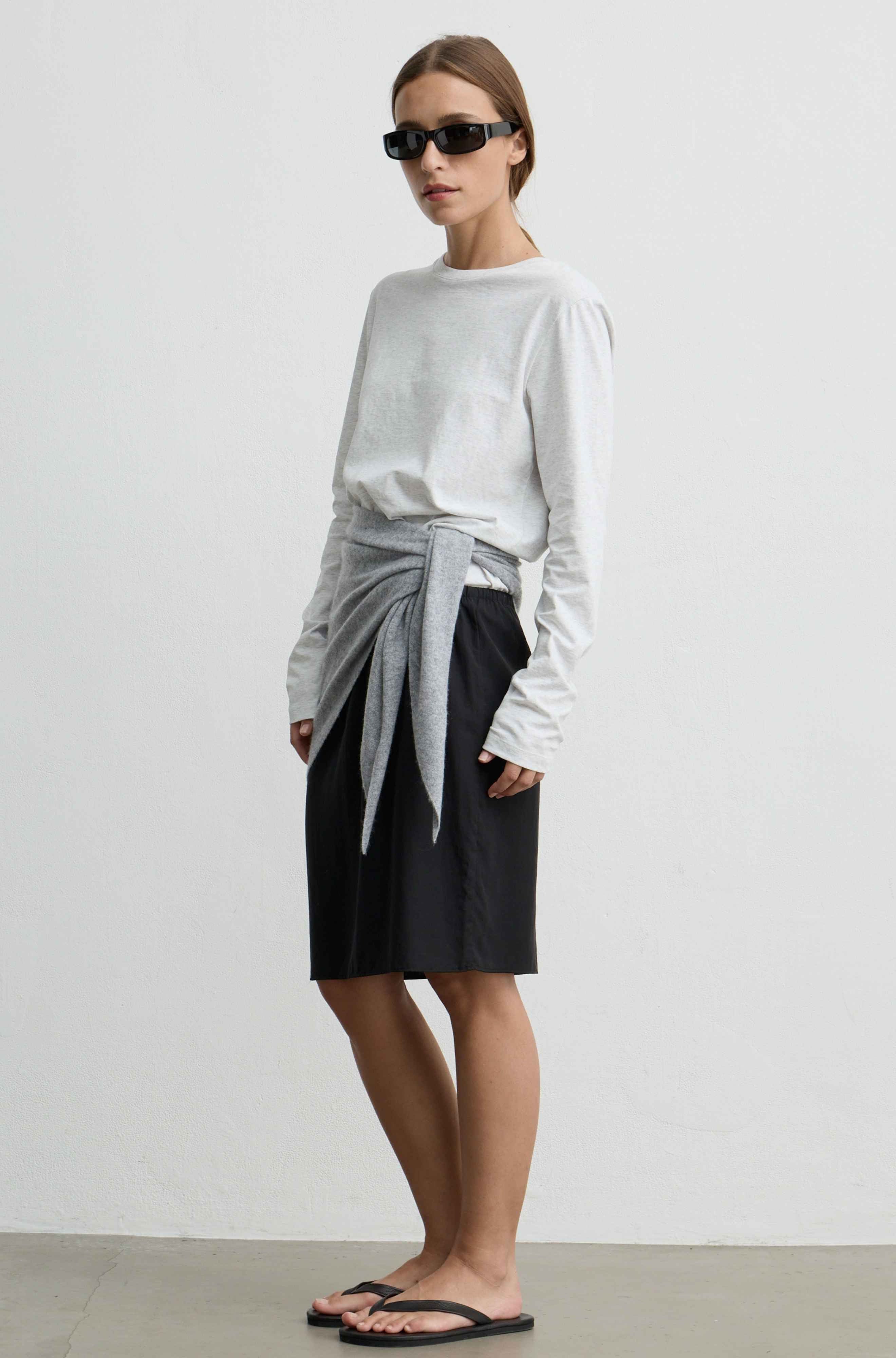 A model wearing a light gray long-sleeve top with a gray knit tie-waist sash, a black midi skirt, and black flip-flops, standing against a plain light gray wall.