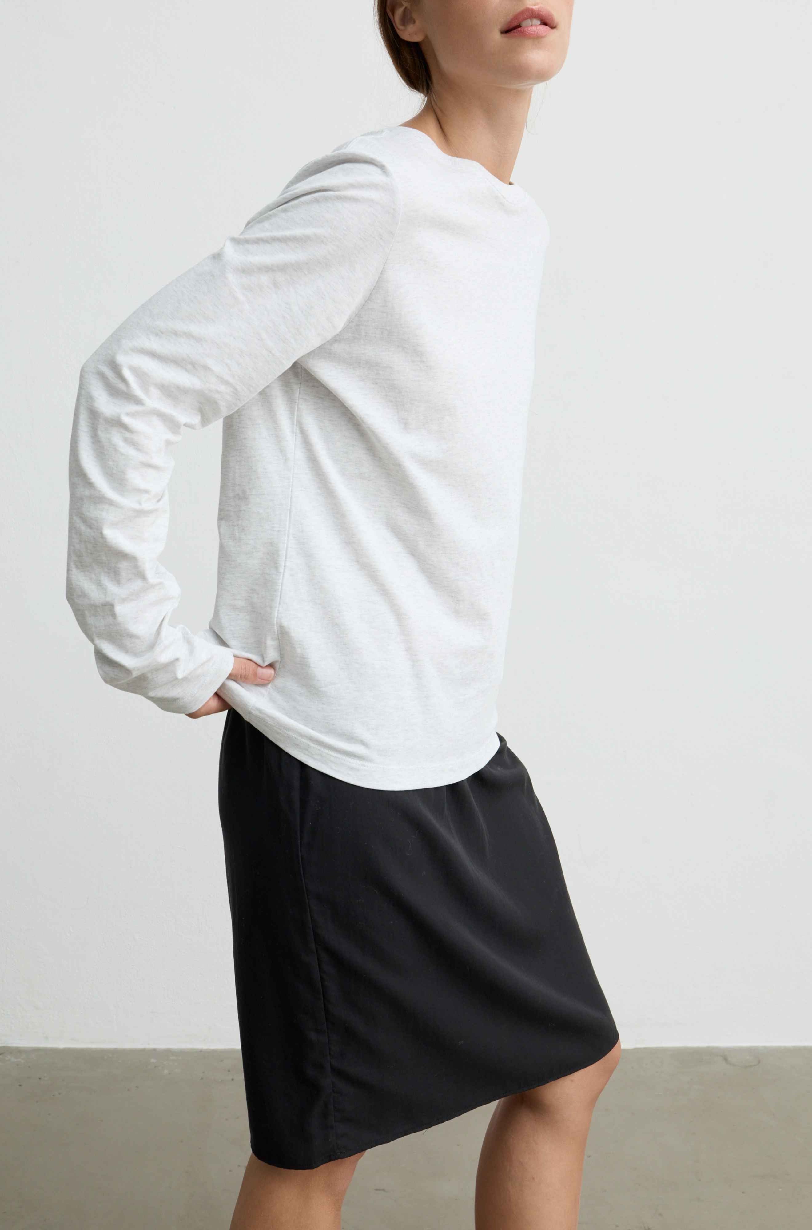 A woman wearing a white long-sleeve crewneck top and a black skirt, shown from the side against a plain light gray wall.