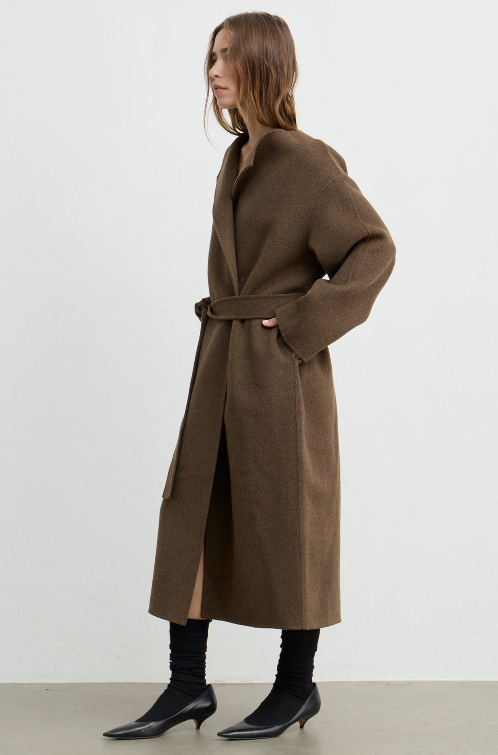 Woman wearing a dark brown wool wrap coat with a tie belt, black heels, and holding a black bag against a white wall