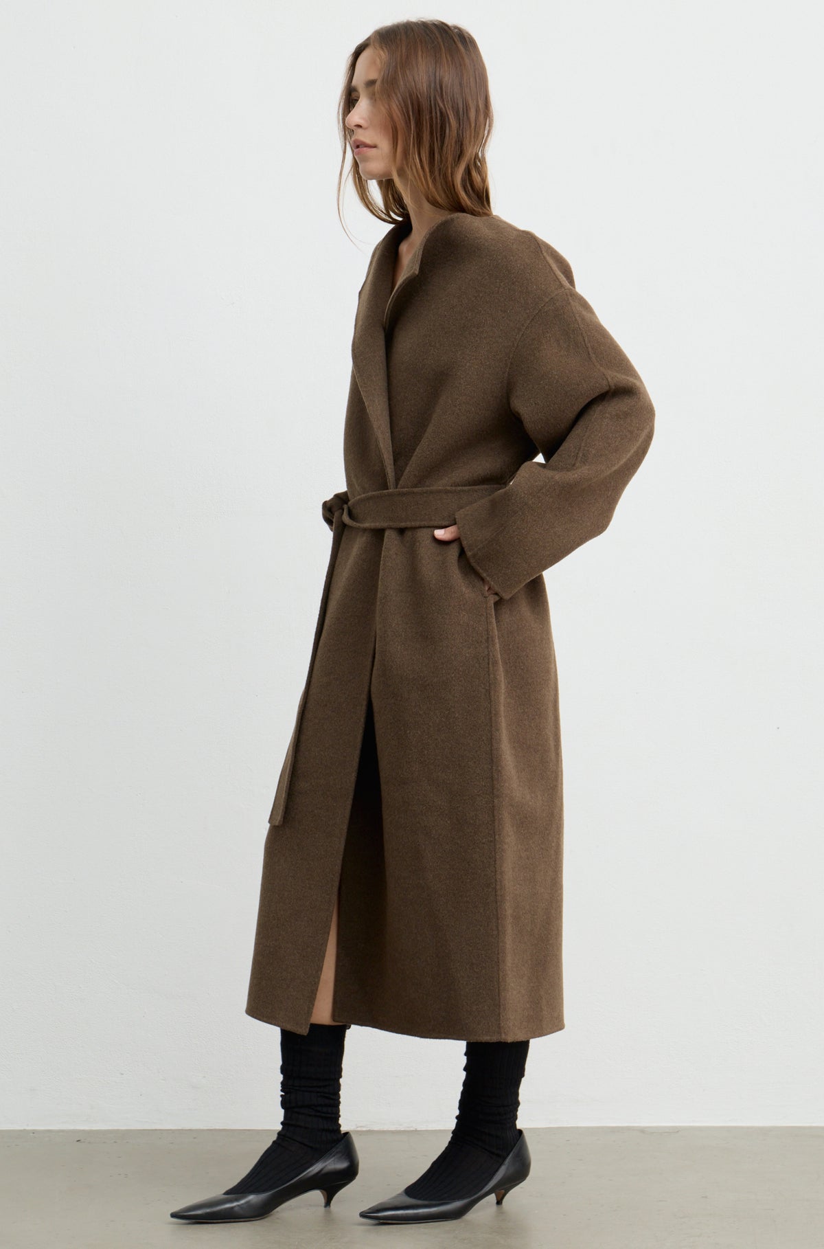Woman wearing a dark brown wool wrap coat with a tie belt, black heels, and holding a black bag against a white wall