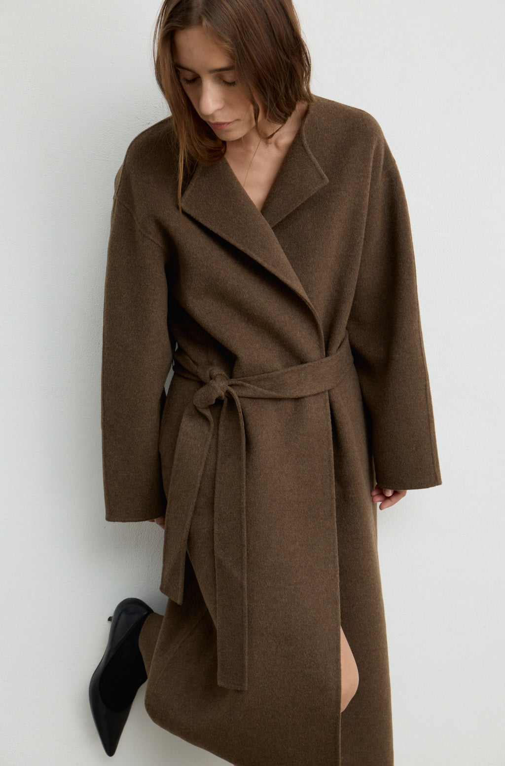 Woman wearing a dark brown wool wrap coat with a tie belt, black heels, and holding a black bag against a white wall