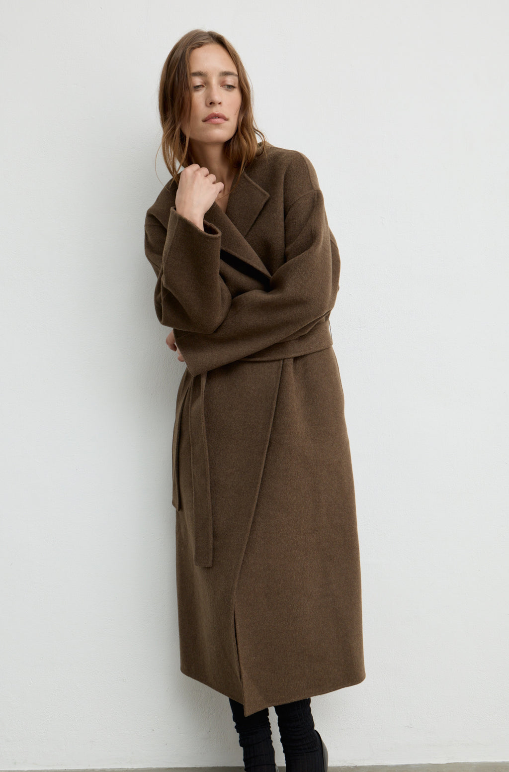 Woman wearing a dark brown wool wrap coat with a tie belt against a white wall