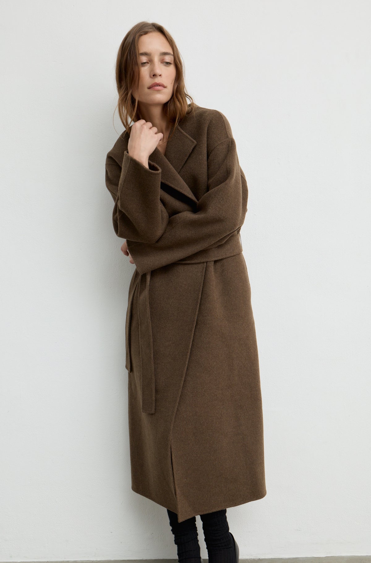 Woman wearing a dark brown wool wrap coat with a tie belt against a white wall