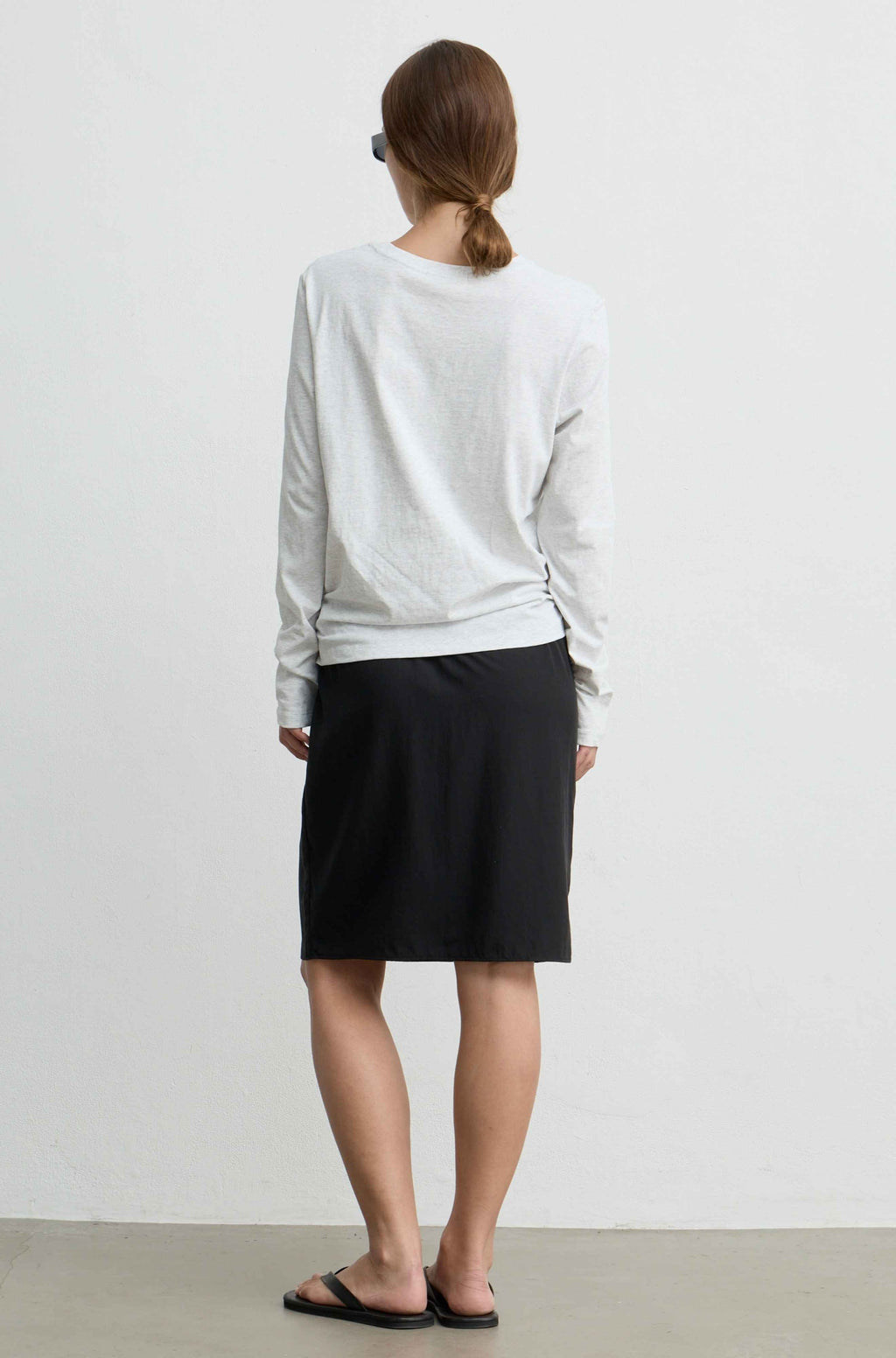 A woman viewed from behind wearing a light gray long-sleeve top and a black knee-length skirt, standing against a plain off-white wall.