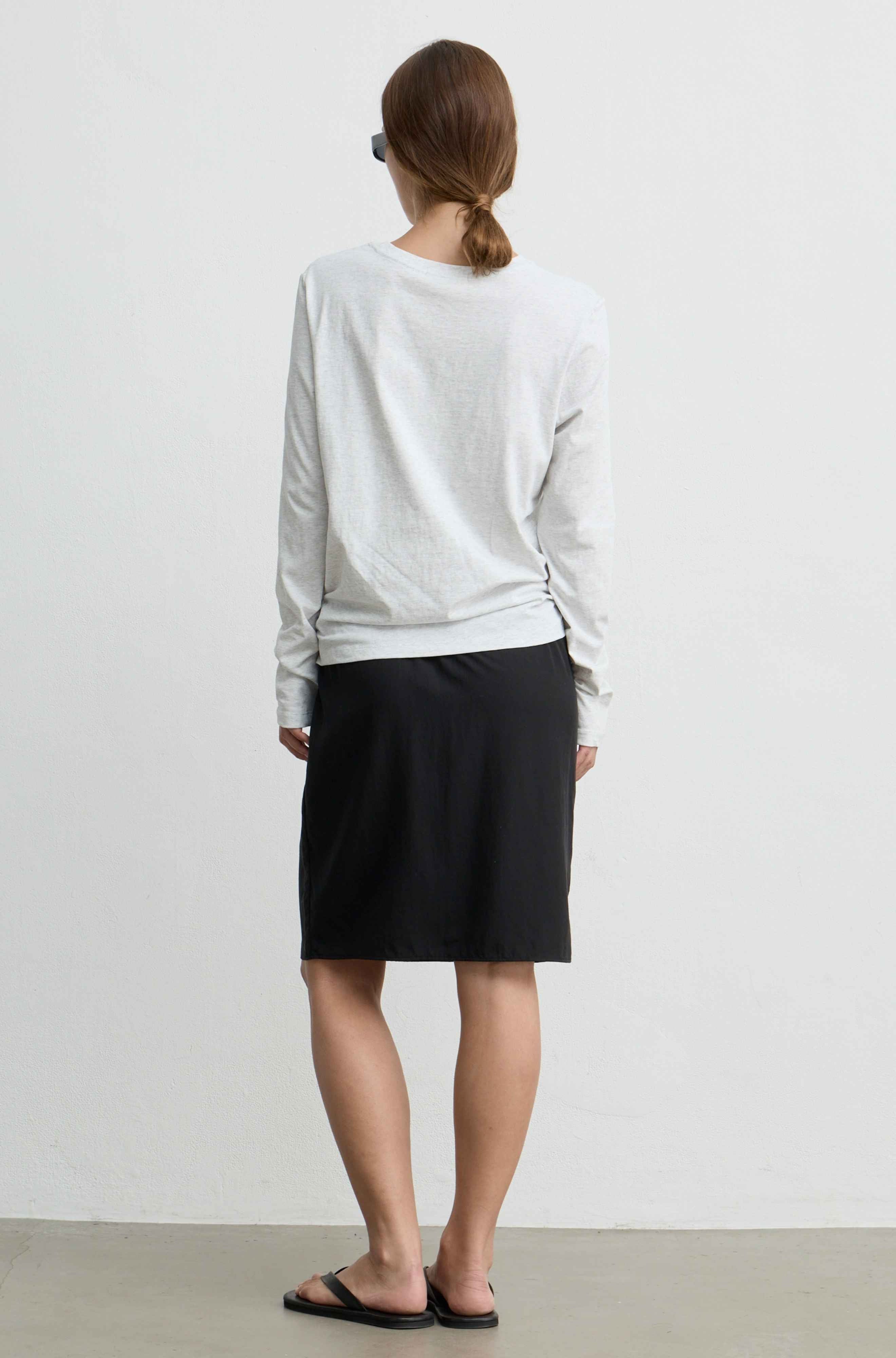 A woman viewed from behind wearing a light gray long-sleeve top and a black knee-length skirt, standing against a plain off-white wall.