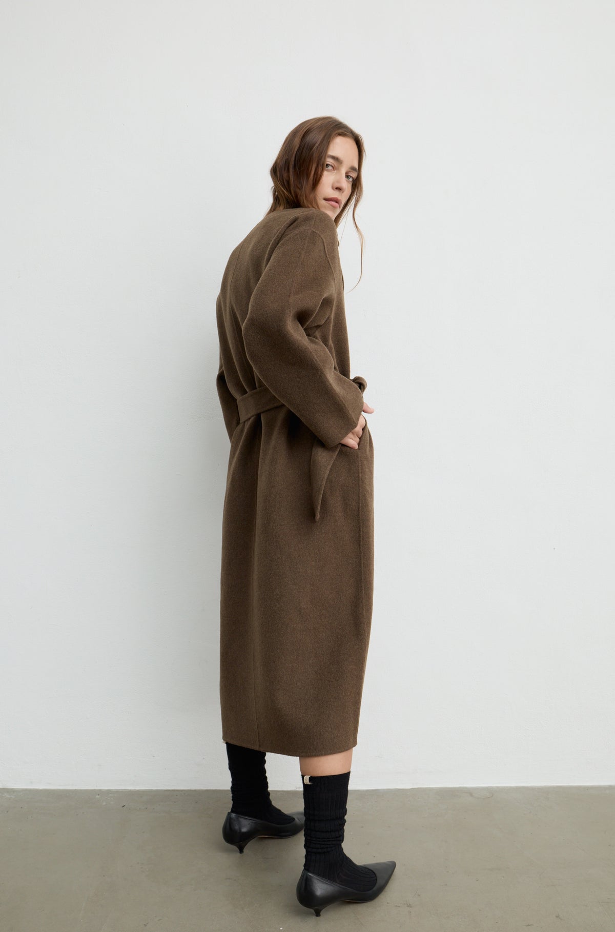 Woman wearing a dark brown wool wrap coat with a tie belt, black heels against a white wall