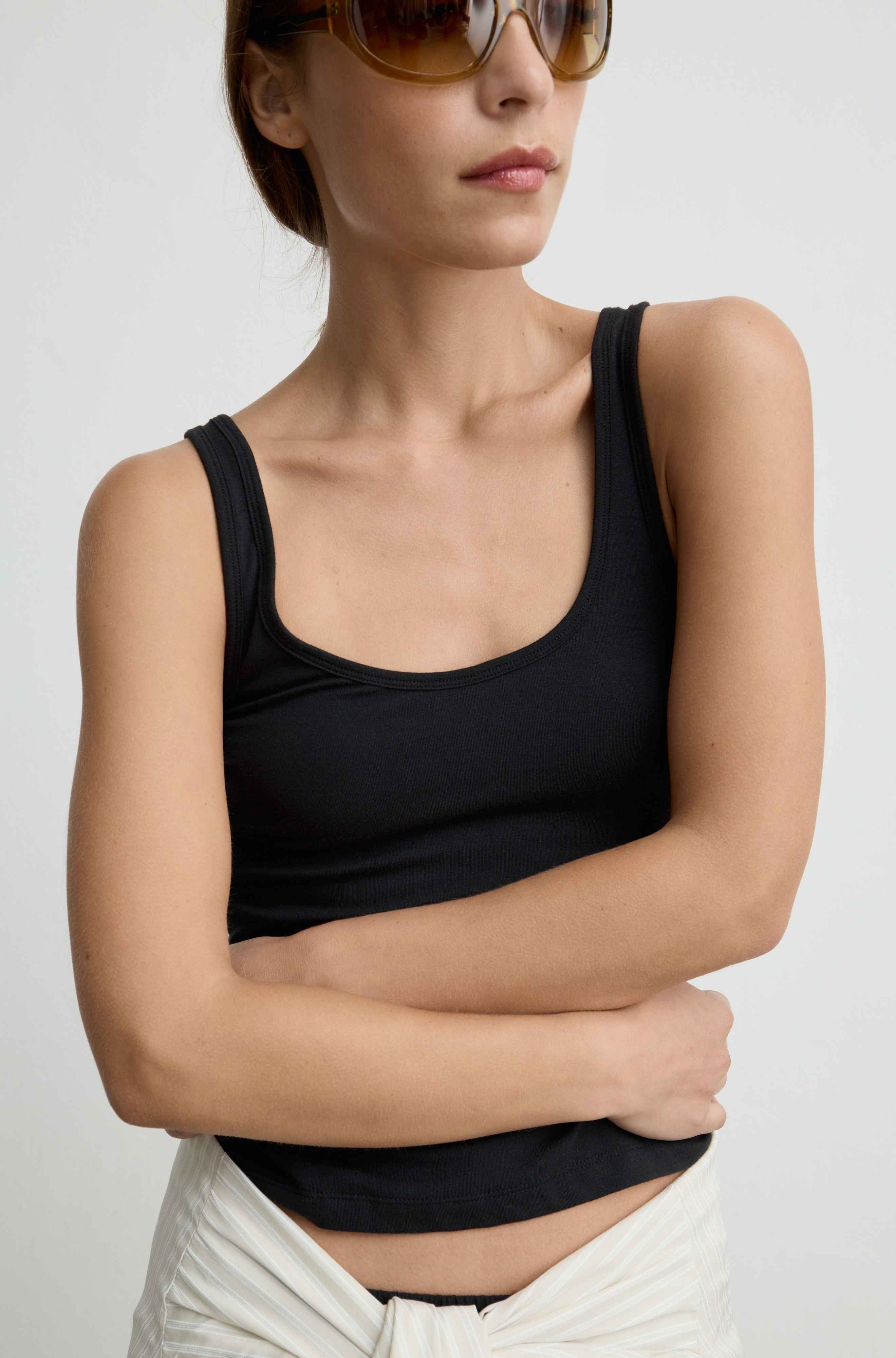 A woman wearing a black scoop-neck tank top and cream-colored pants, photographed against a plain light gray background.