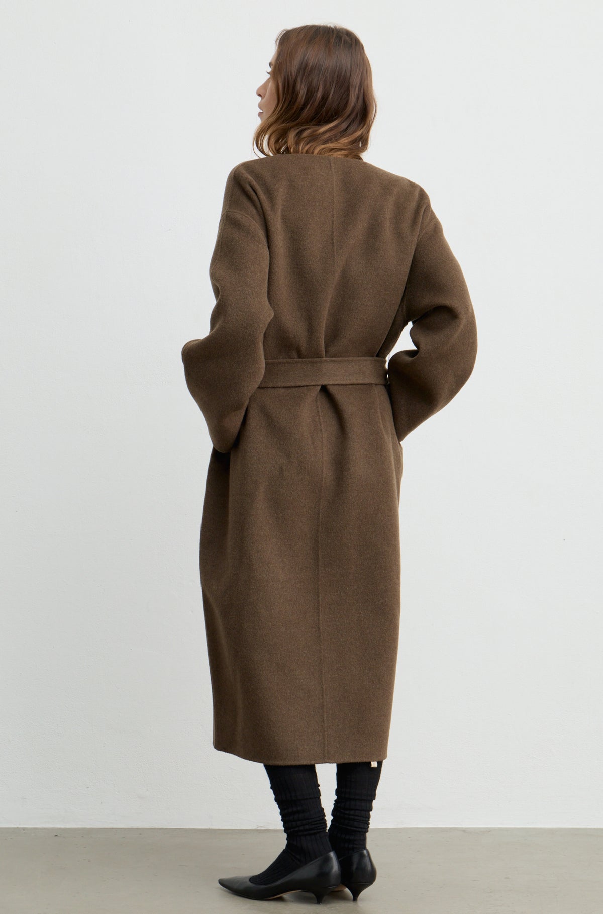 Woman wearing a dark brown wool wrap coat with a tie belt against a white wall