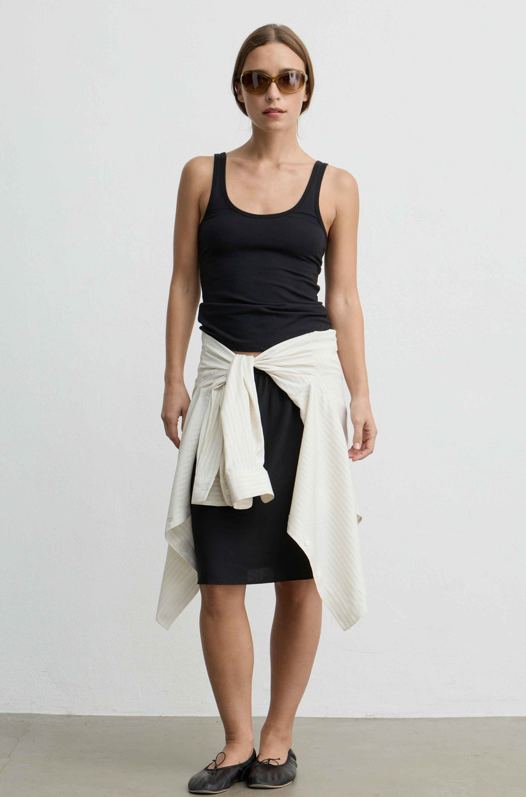 A woman modeling a black tank top, black skirt, and a cream-colored knit sweater tied around her waist, paired with dark flat shoes and sunglasses, against a plain white studio background.