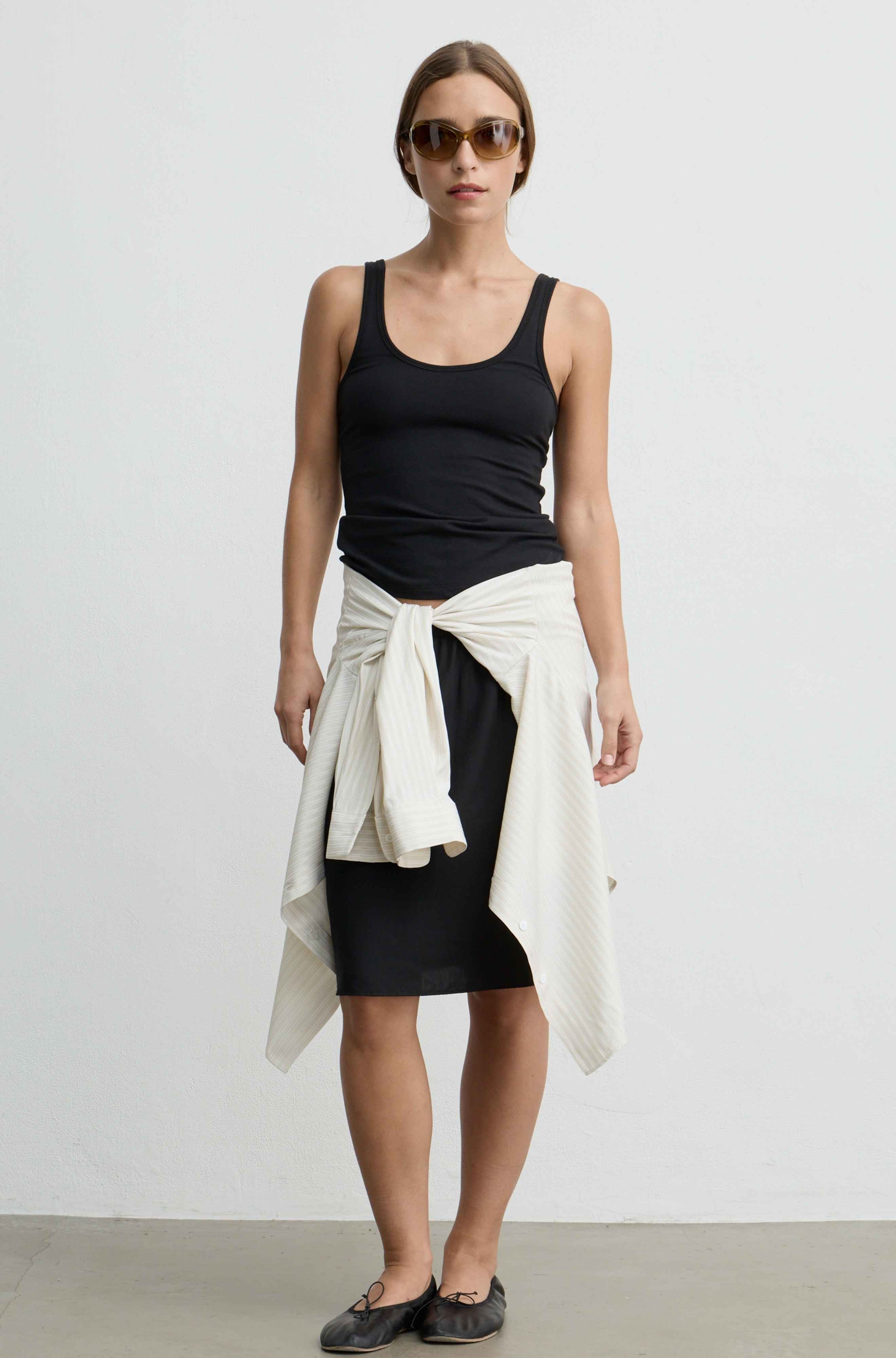 A woman modeling a black tank top, black skirt, and a cream-colored knit sweater tied around her waist, paired with dark flat shoes and sunglasses, against a plain white studio background.