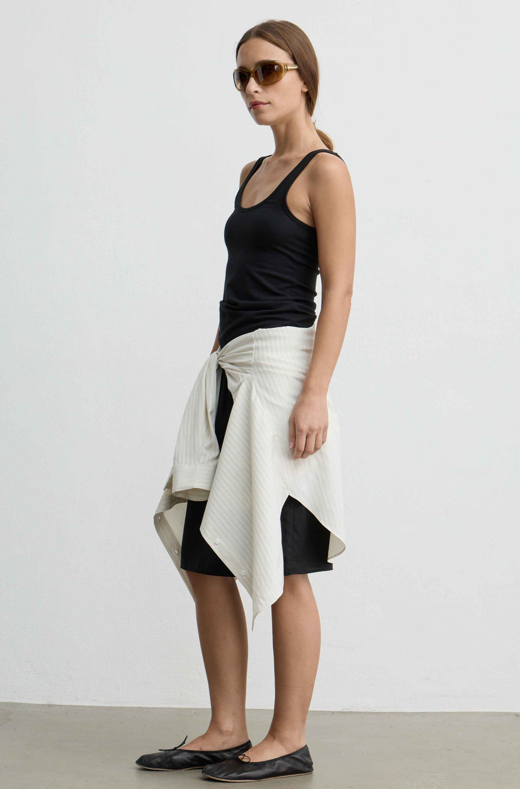A woman modeling a black ribbed tank top, black shorts, and a cream-colored textured wrap skirt tied at the waist, styled with sunglasses and black flats against a plain white studio background.