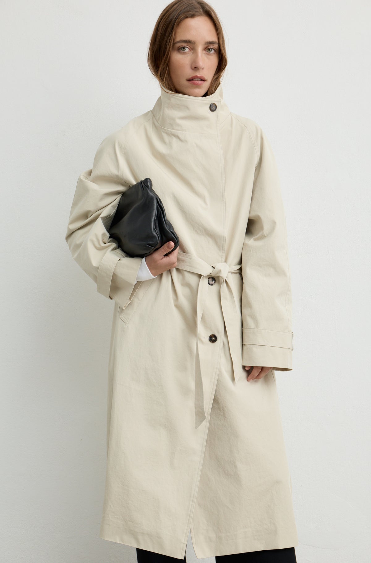 Woman wearing a beige trench coat with a high neckline and button up front, holding a black handbag against a light grey wall