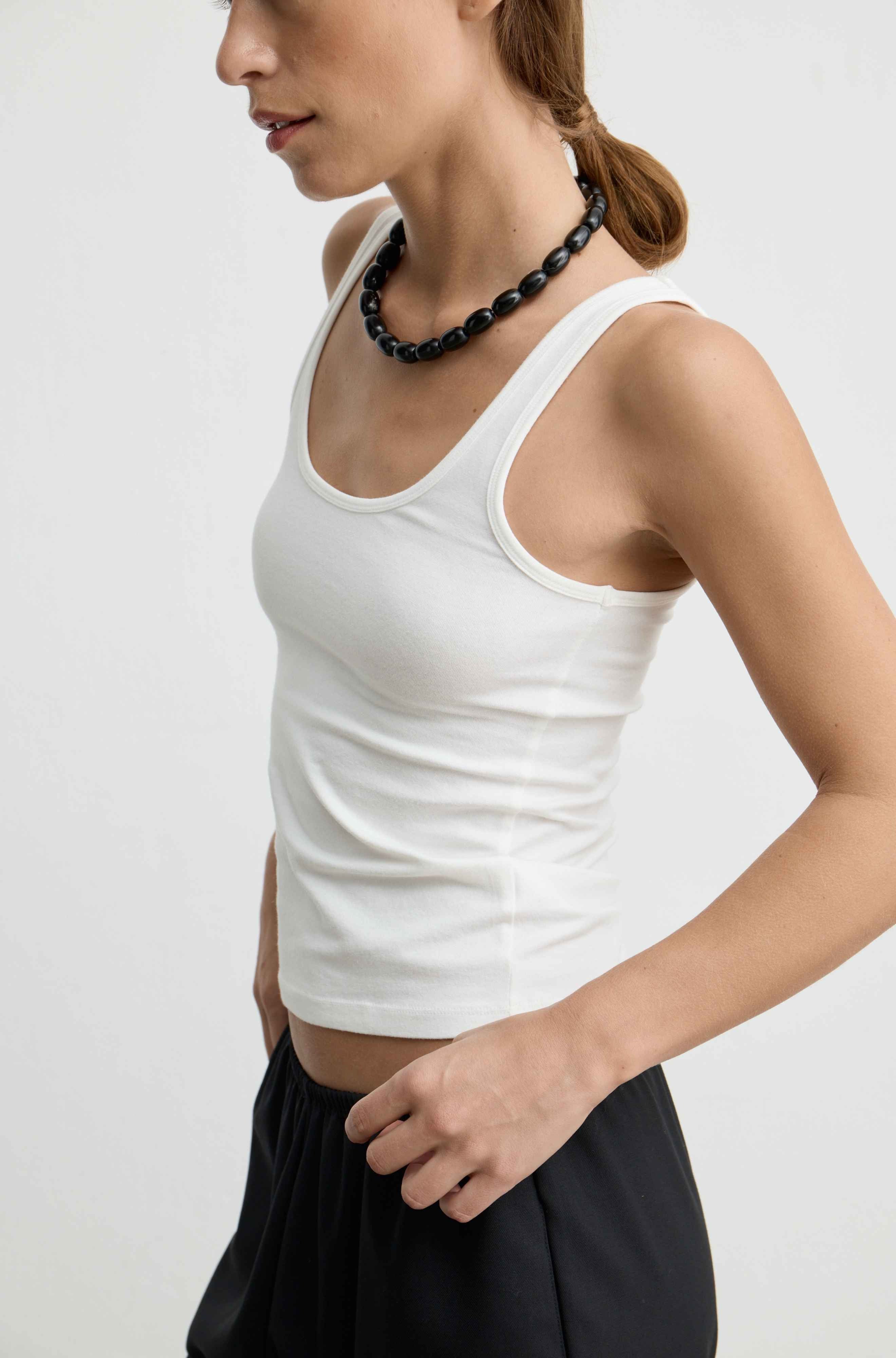 A woman wearing a fitted white tank top and black skirt, shown from the side against a plain light background.