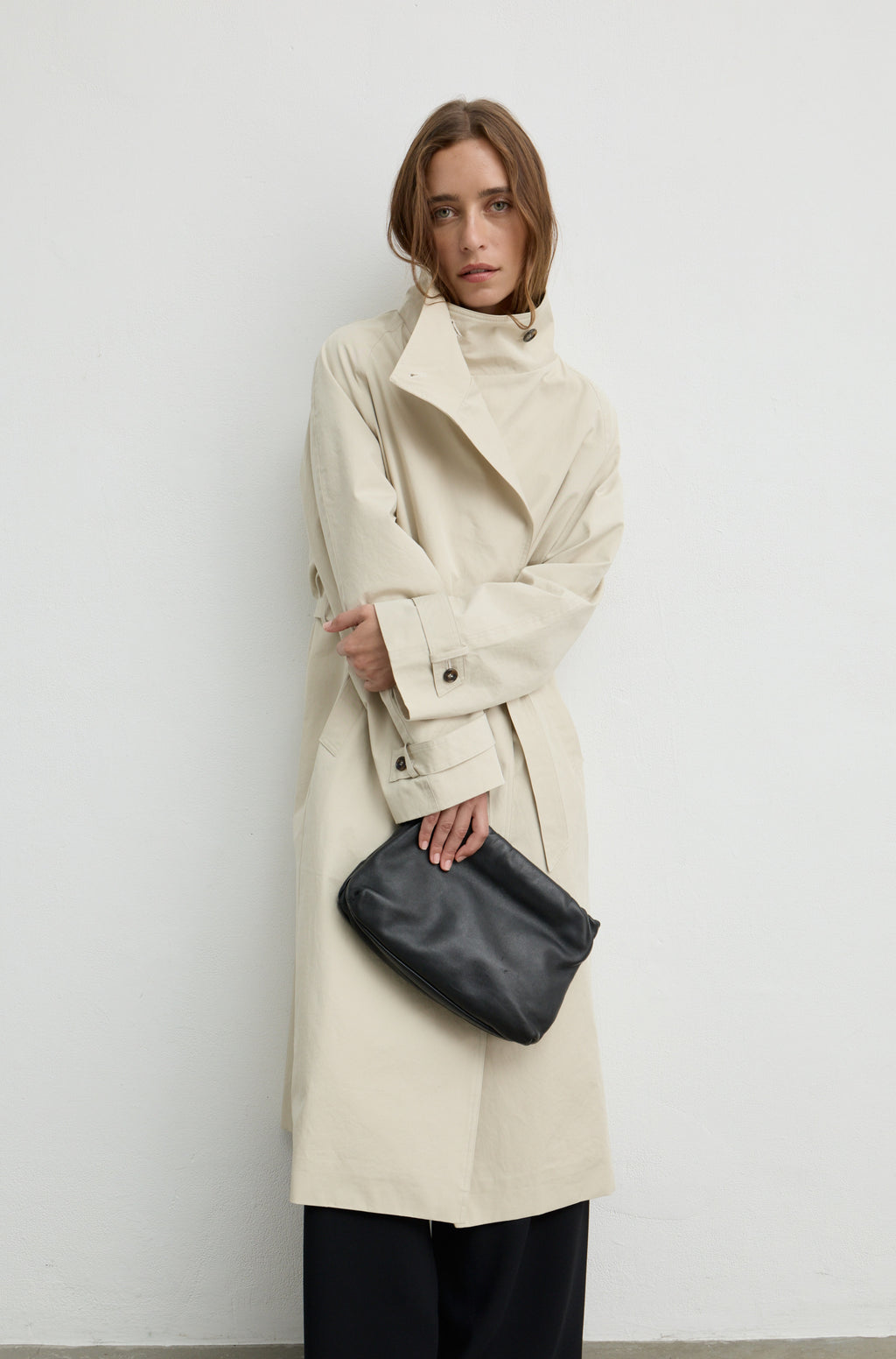 Woman wearing a beige trench coat with a high neckline and button up front, holding a black handbag against a light grey wall