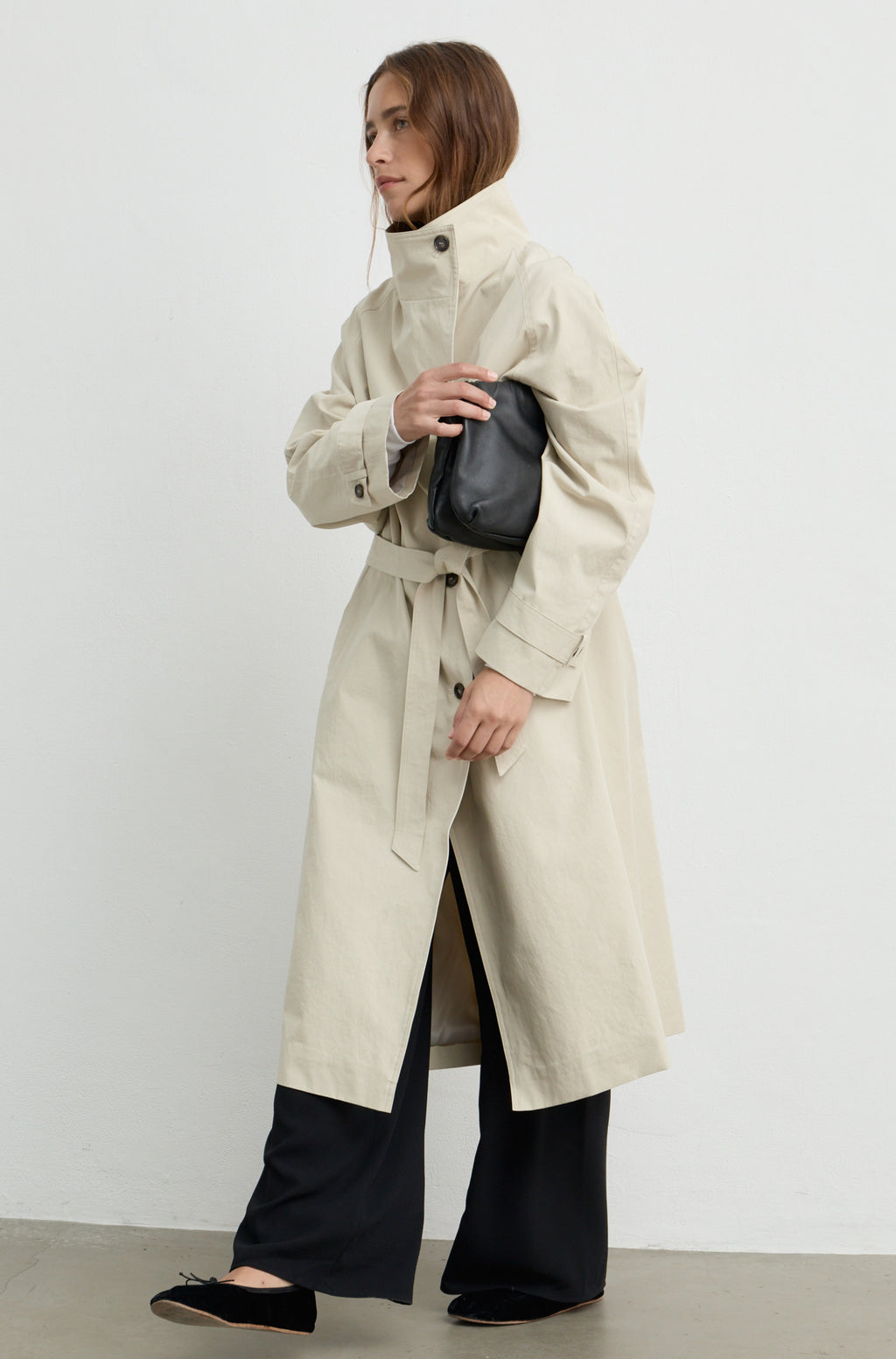 Woman wearing a beige trench coat with a high neckline and button up front, holding a black handbag against a light grey wall