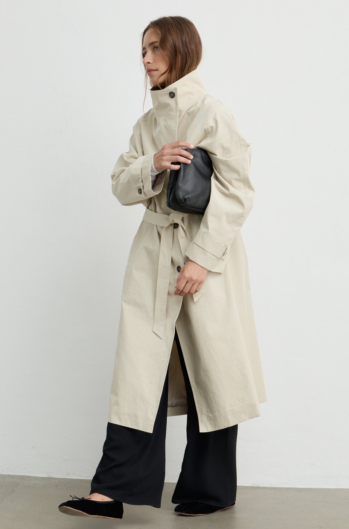 Woman wearing a beige trench coat with a high neckline and button up front, holding a black handbag against a light grey wall