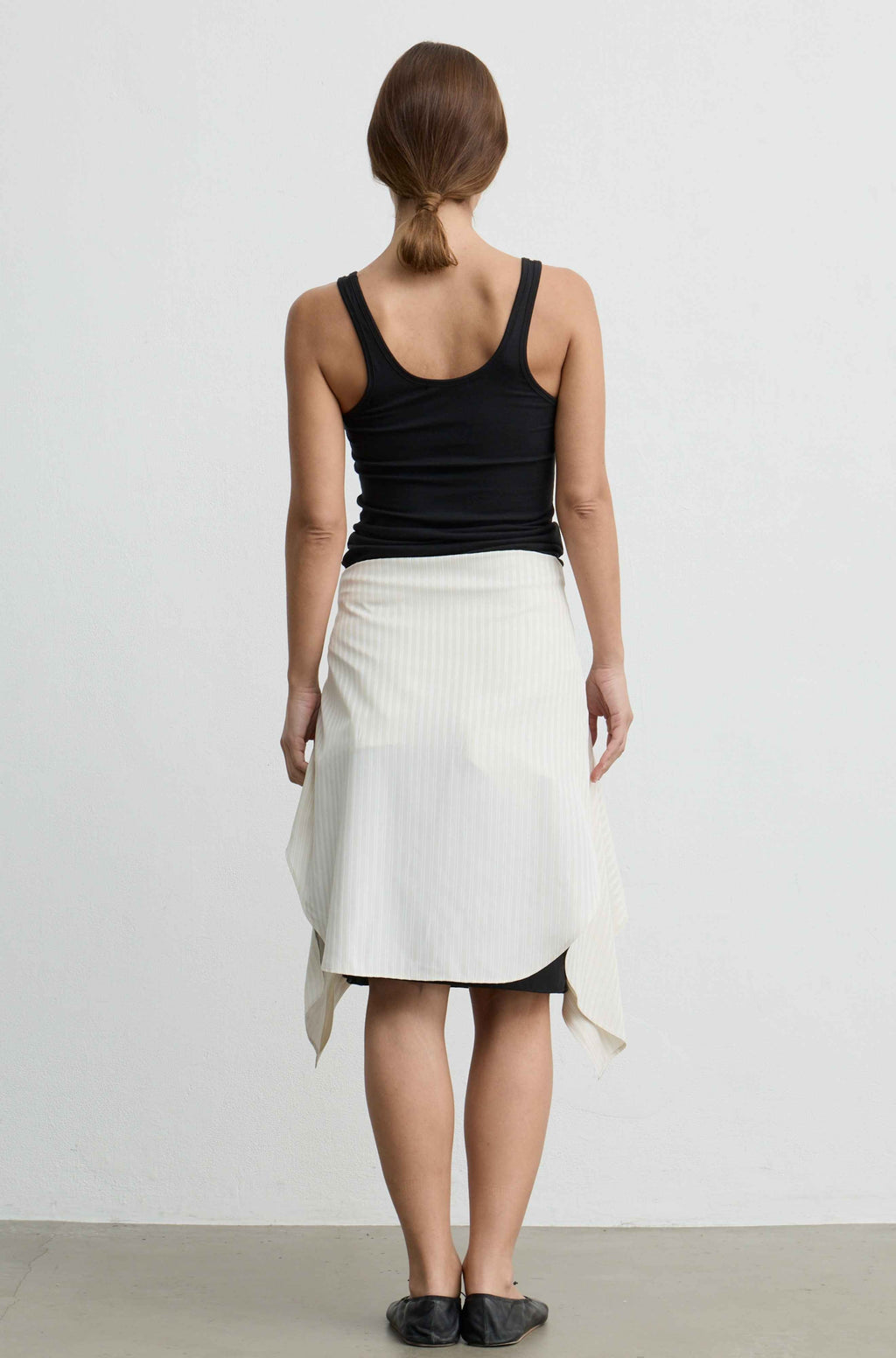 A woman shown from the back wearing a black tank top and a white wrap-style midi skirt with a black underlayer visible at the hem, standing against a plain light gray wall.