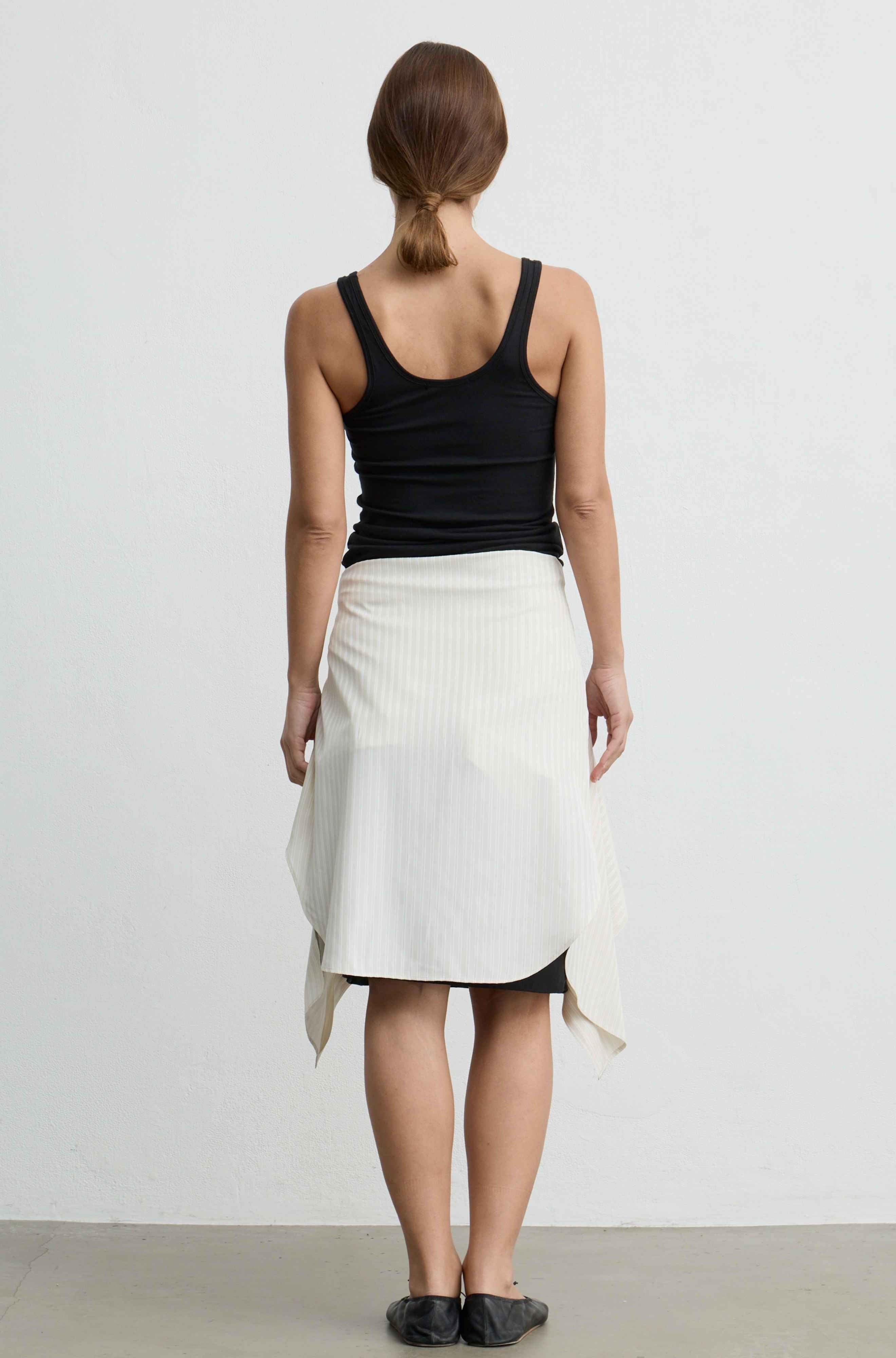 A woman shown from the back wearing a black tank top and a white wrap-style midi skirt with a black underlayer visible at the hem, standing against a plain light gray wall.