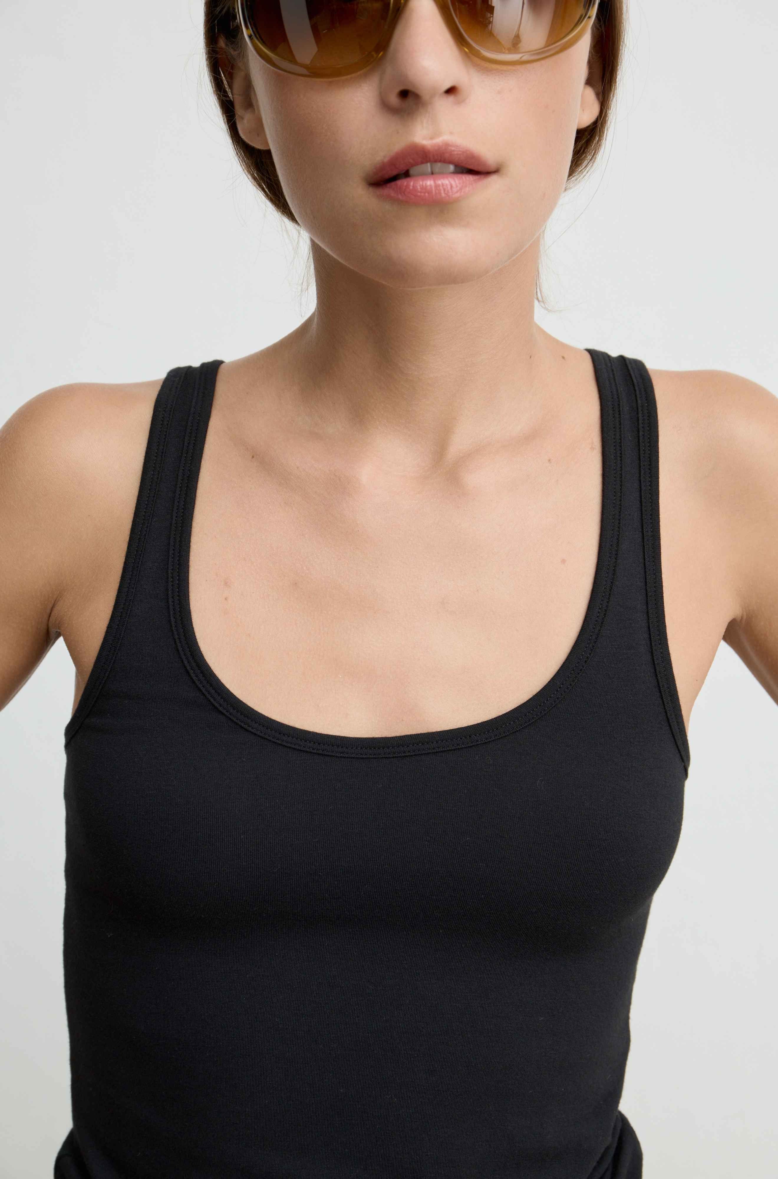 A close-up of a model wearing a black scoop-neck tank top against a light gray background.