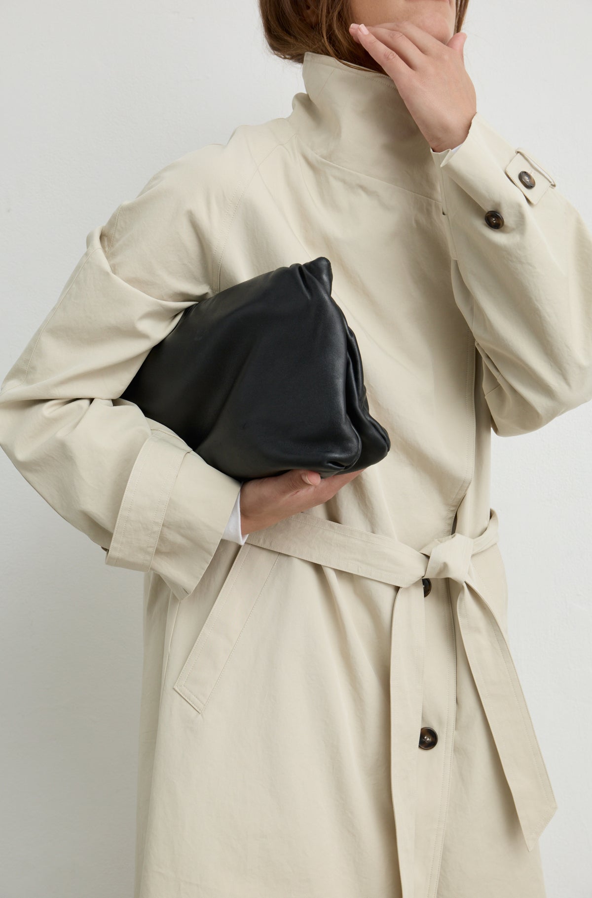 Woman wearing a beige trench coat with a high neckline and button up front, holding a black handbag against a light grey wall