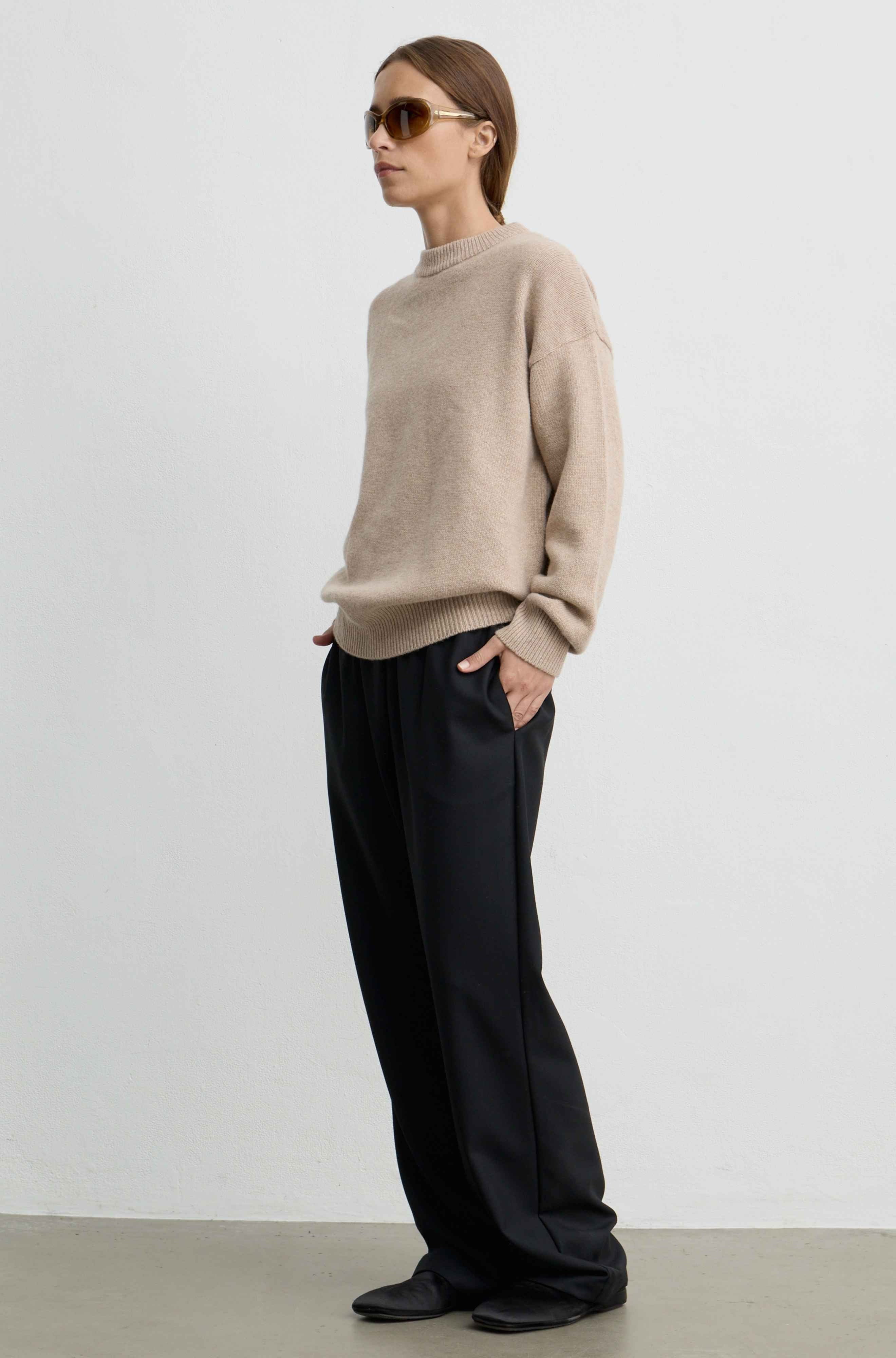 A woman modeling a light beige knit sweater and black wide-leg trousers, standing in profile against a plain light gray studio wall.