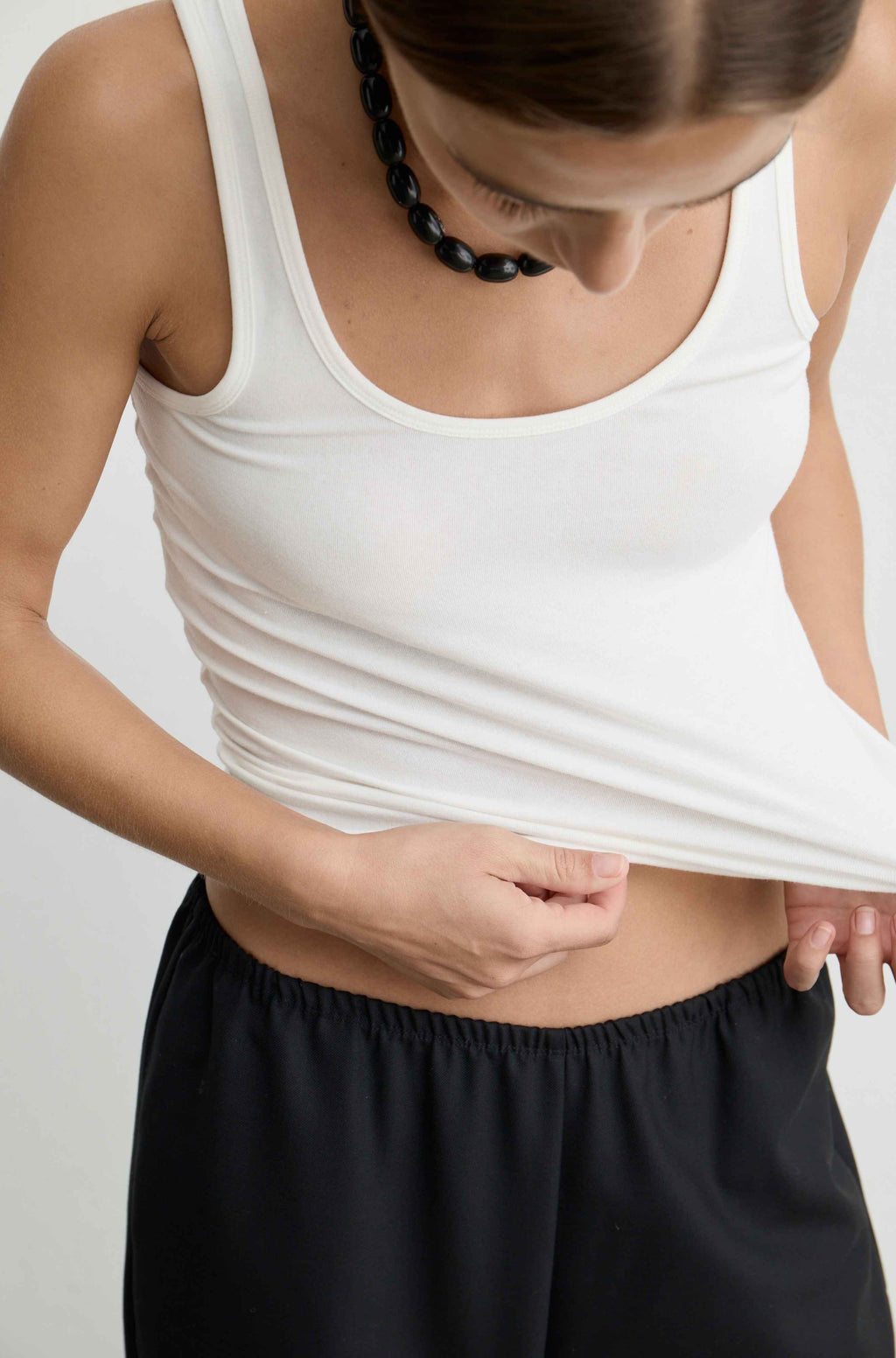A close-up of a woman wearing a white tank top and black shorts, lifting the hem of the tank top to show her midriff. She is wearing a chunky black beaded necklace and looking down.
