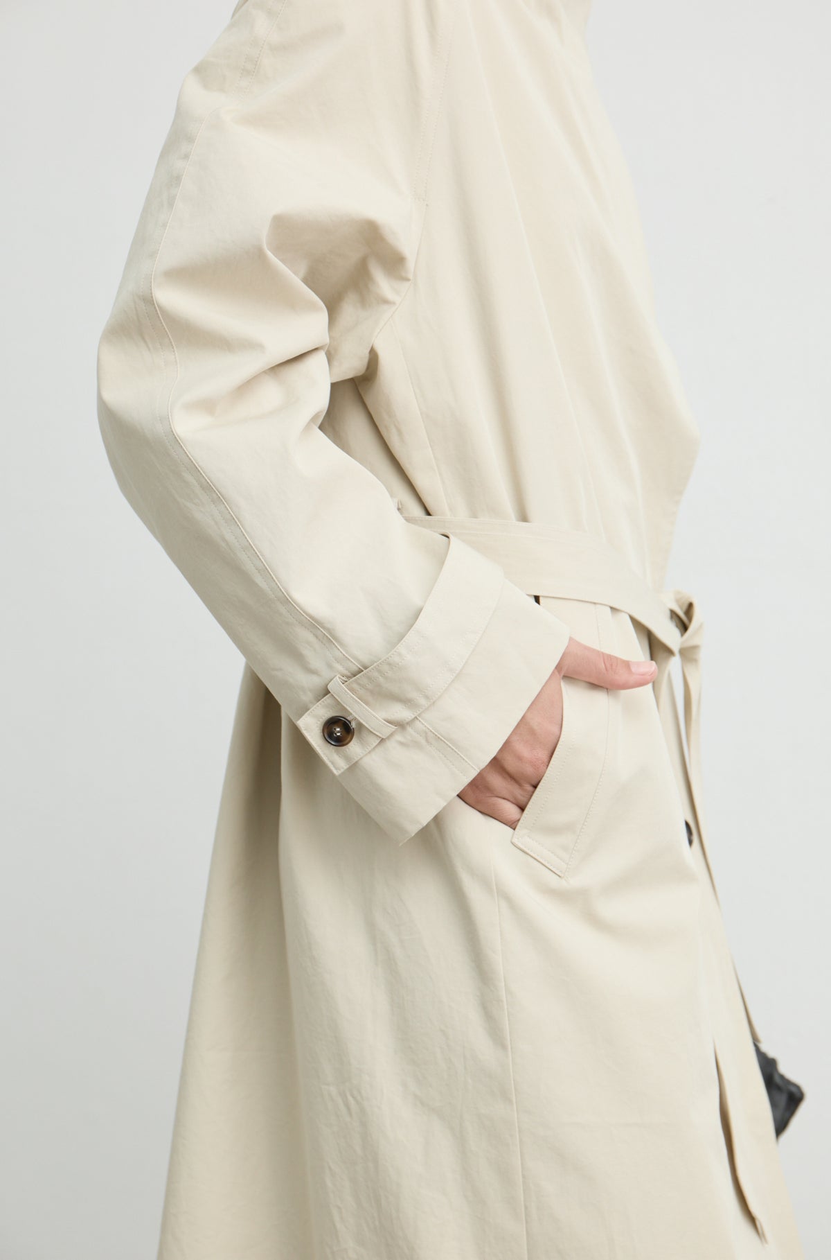 Woman wearing a beige trench coat with her hand in the side pocket, against a light grey wall