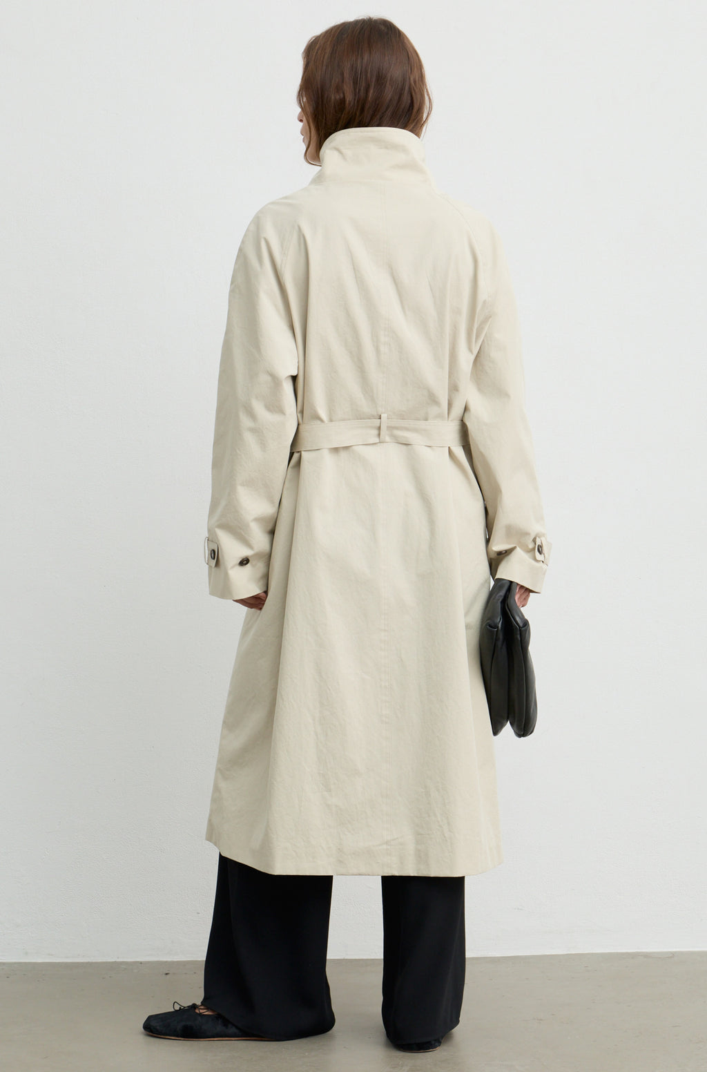 Woman wearing a beige trench coat with a high neckline and button up front, holding a black handbag against a light grey wall
