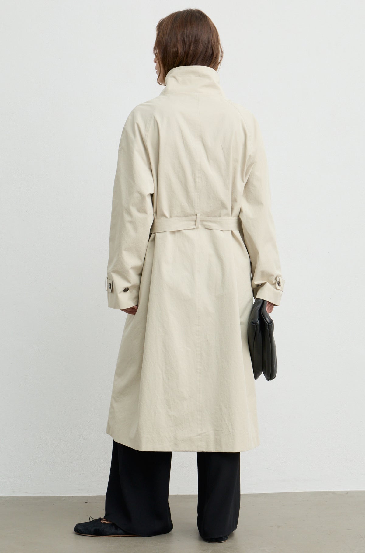 Woman wearing a beige trench coat with a high neckline and button up front, holding a black handbag against a light grey wall