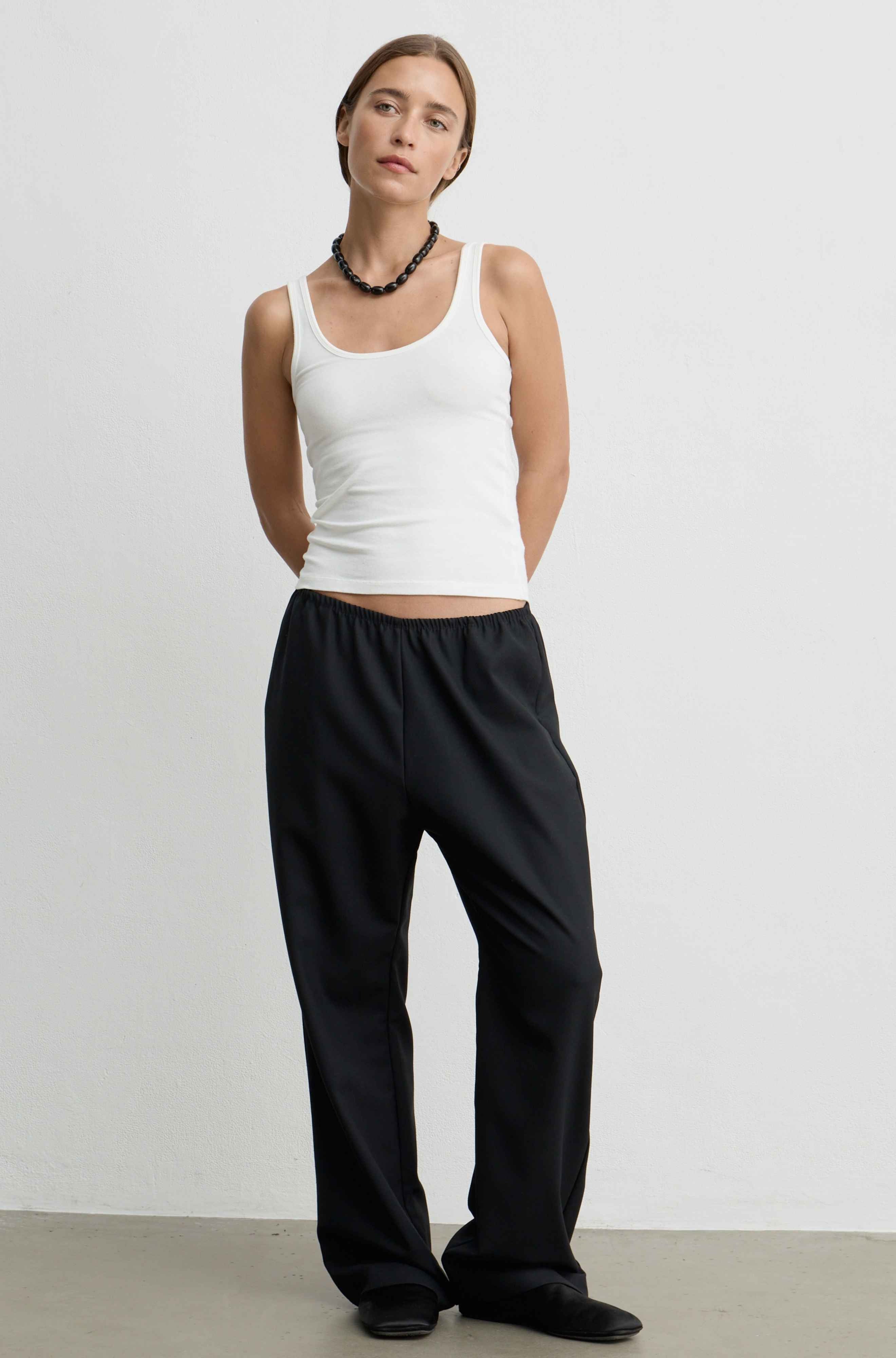 A woman modeling a white tank top and black wide-leg pants against a plain light gray studio wall.