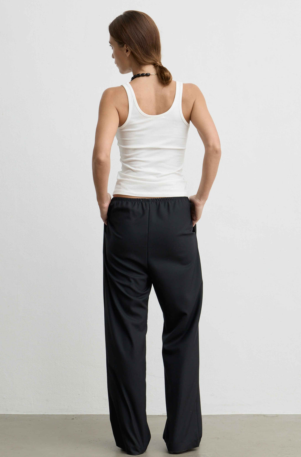 A woman shown from the back wearing a white tank top and black wide-leg pants against a plain light gray studio background.