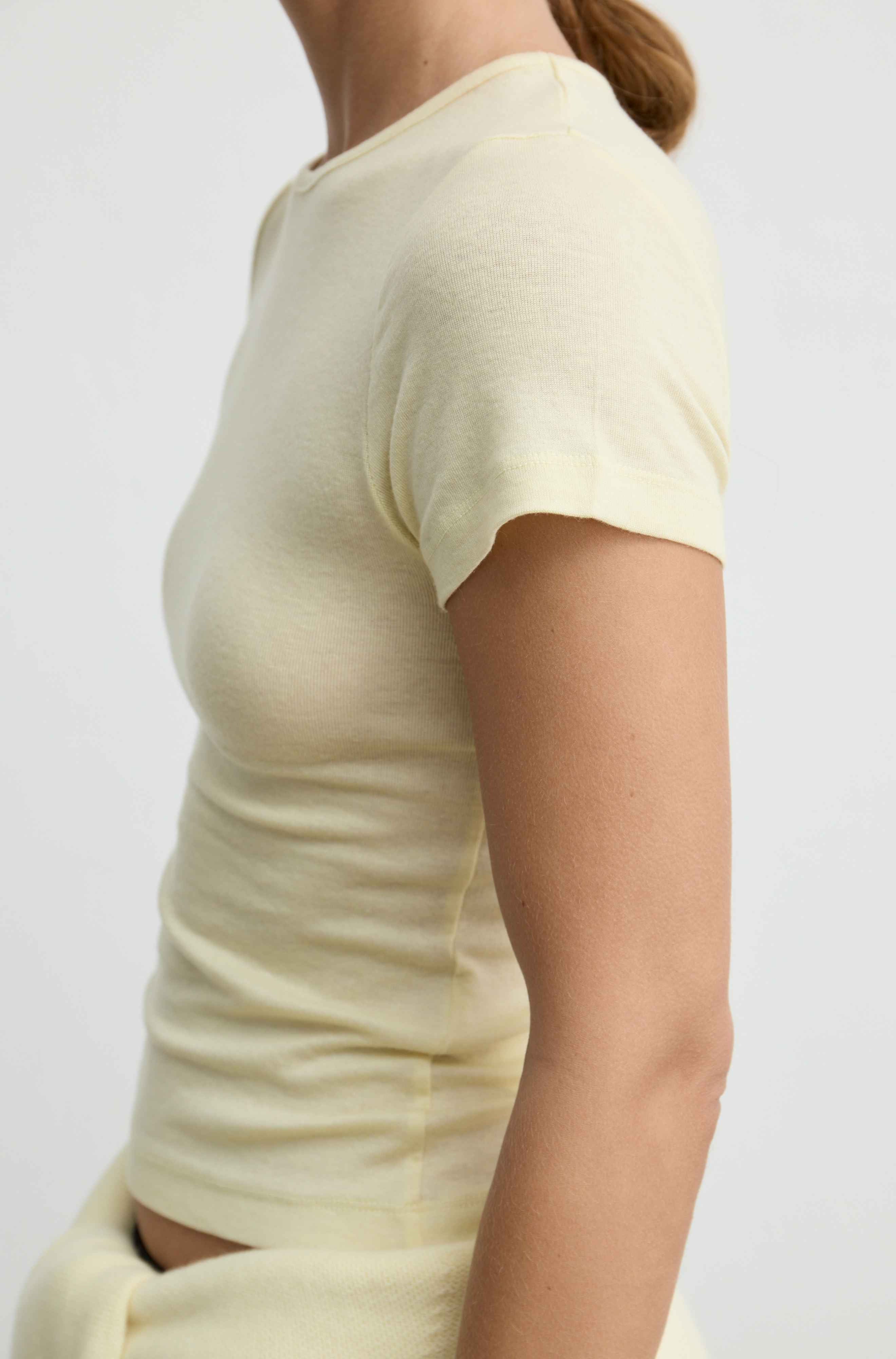 A close-up side view of a person wearing a pale butter yellow short-sleeve ribbed knit top against a plain light background.