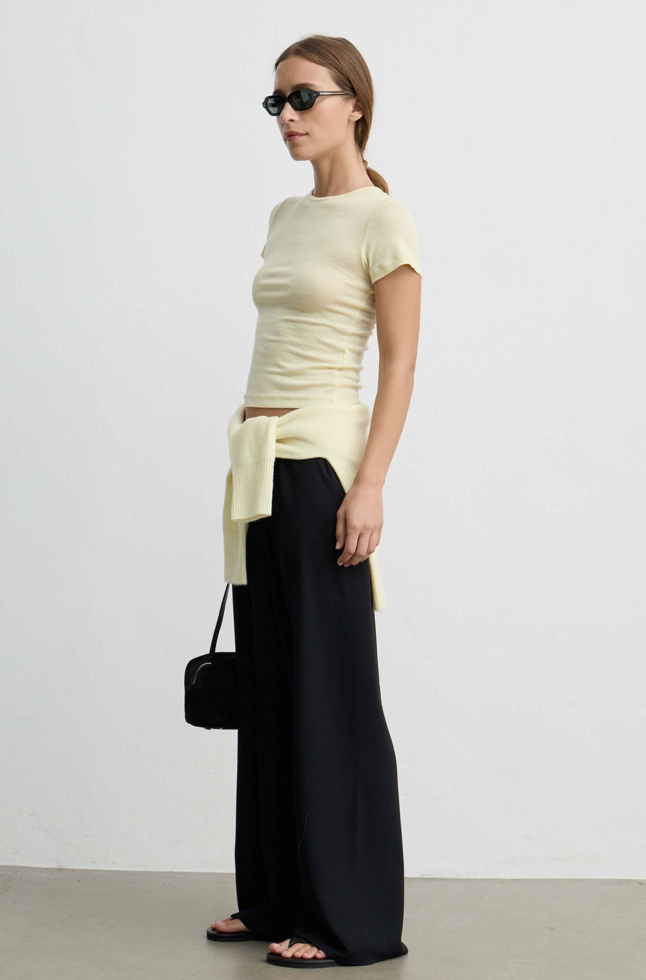A woman modeling a pale yellow ribbed short-sleeve top with a side tie detail, paired with black wide-leg trousers, black sunglasses, and carrying a small black handbag against a plain white studio background.