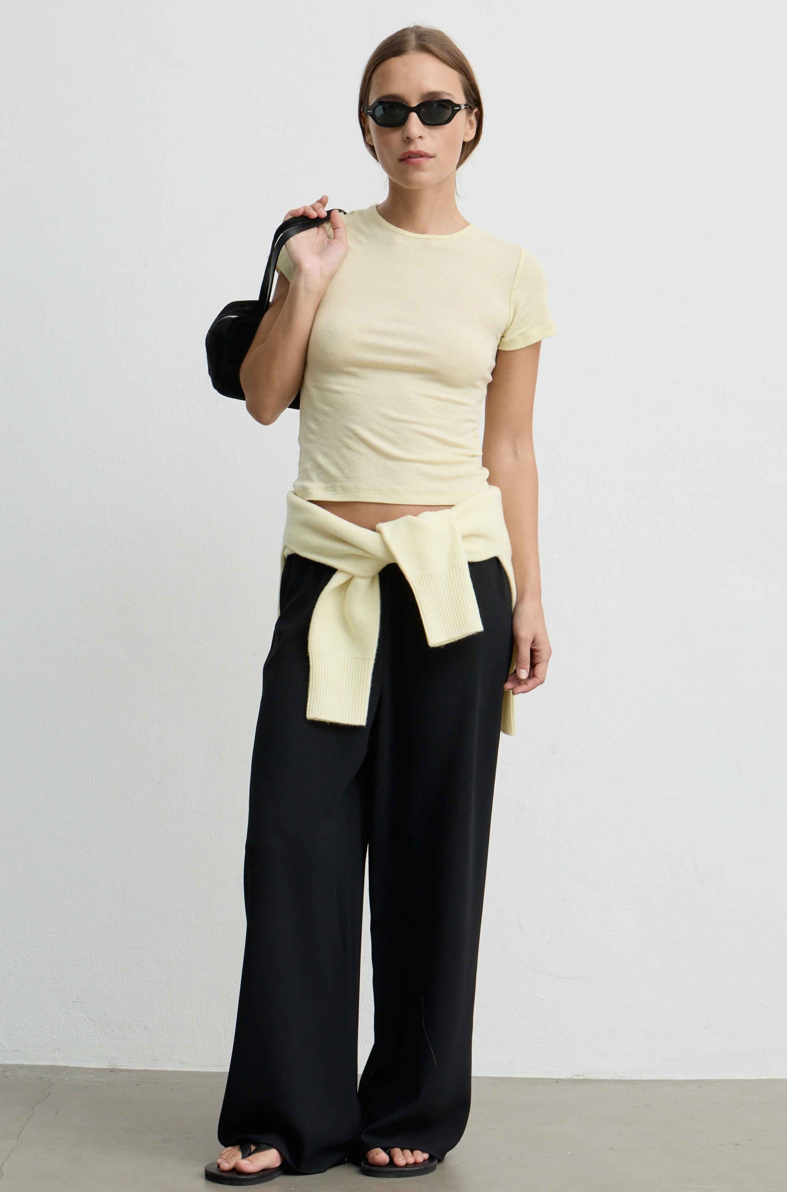 A model stands against a plain white studio wall wearing a pale yellow ribbed short-sleeve crop top with a tied waist sash, black wide-leg pants, black sandals, and a black handbag.