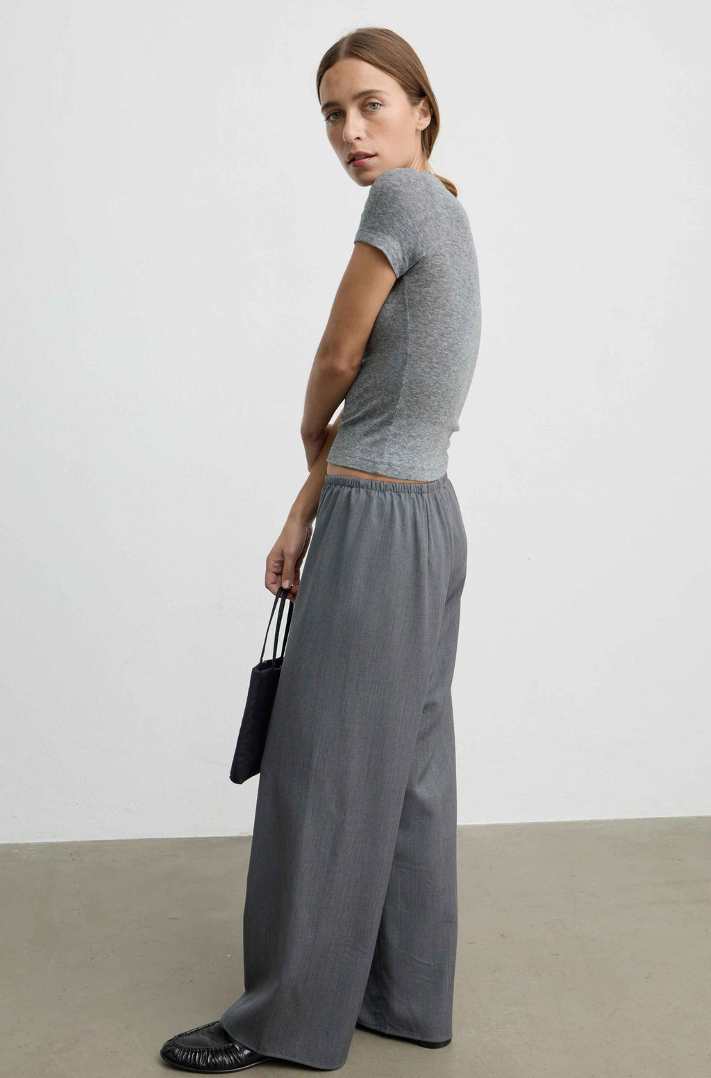 A woman stands in profile wearing a fitted heather gray short-sleeve crop top and matching wide-leg gray trousers, holding a small black handbag against a plain light gray studio background.